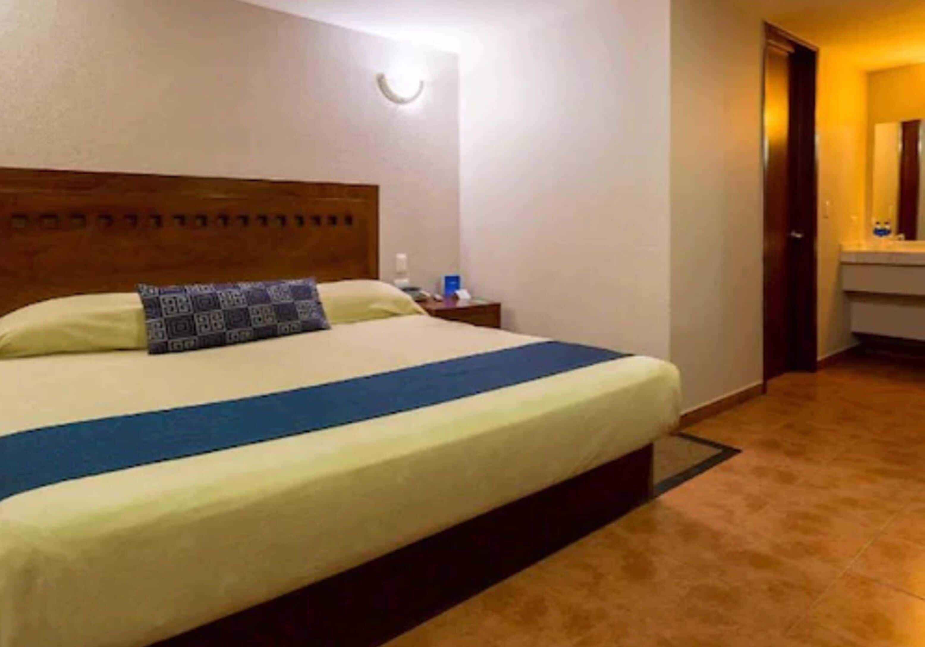 Une chambre d’hôtel avec un lit, une table de chevet, un miroir et une salle de bain visible à travers une porte ouverte.