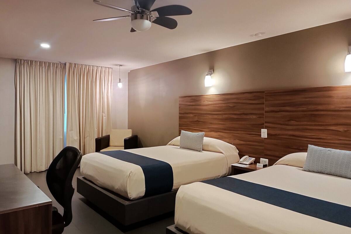 A hotel room with two beds, a desk, a chair, a ceiling fan, and a window with curtains.