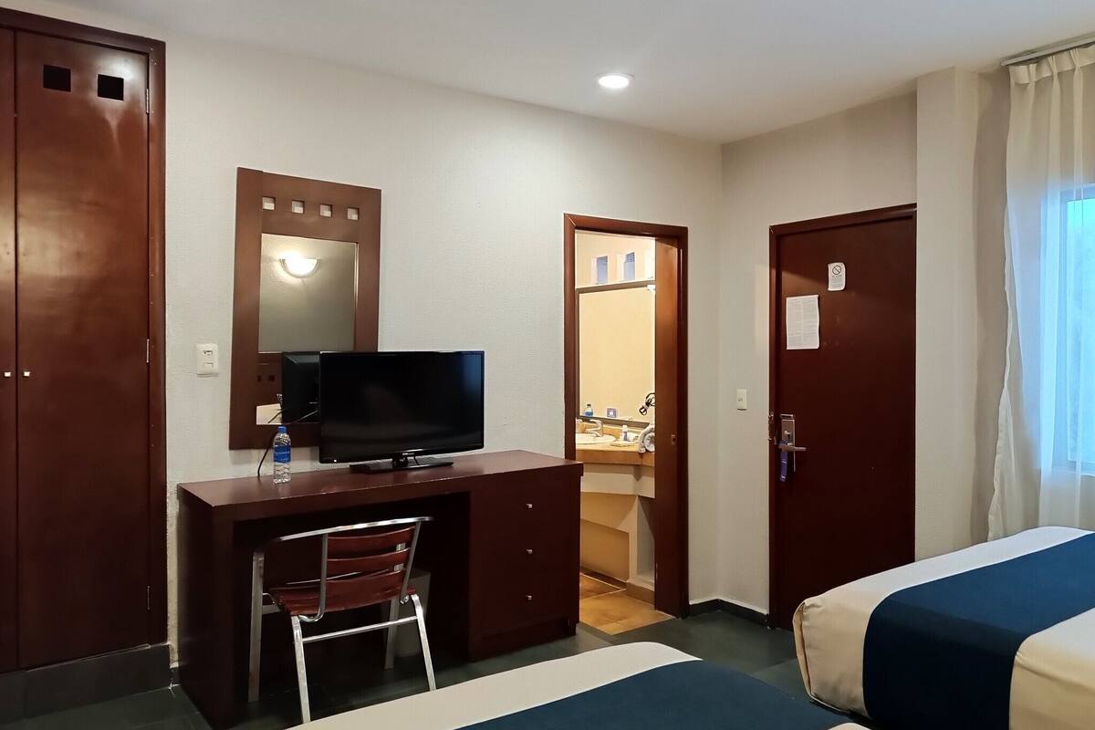 A hotel room with a bed, a desk with a television, a chair, and a bathroom visible through an open door.