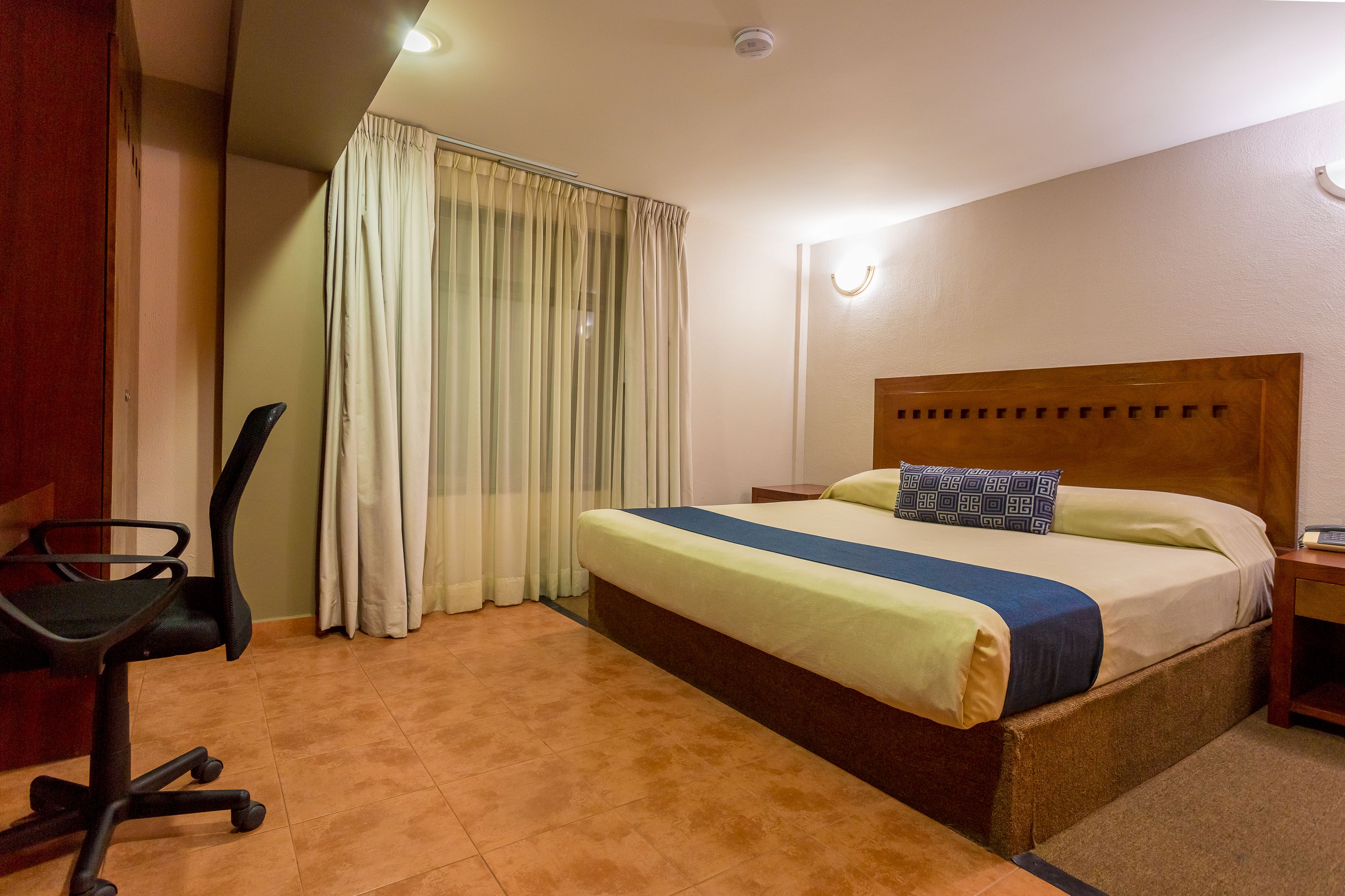 A hotel room with a bed, a desk, a chair, and a window with curtains.