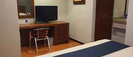 A hotel room with a bed, a desk with a computer, a chair, a mirror, and a wardrobe.