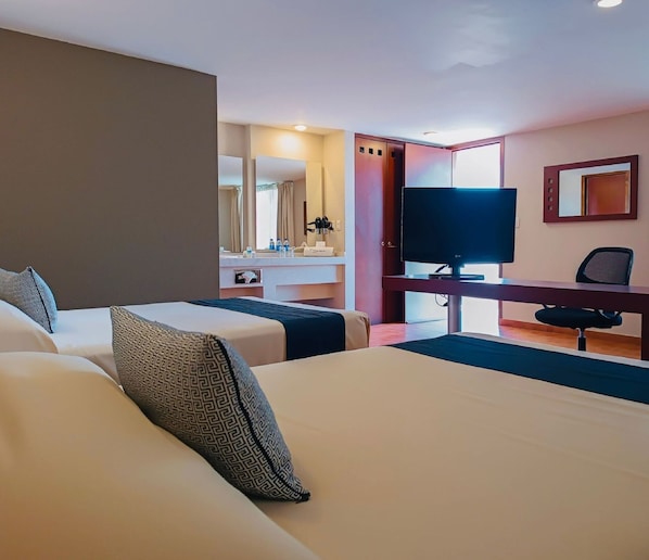 Habitación de hotel con dos camas, un escritorio con televisor y un baño a la vista a través de una puerta abierta.