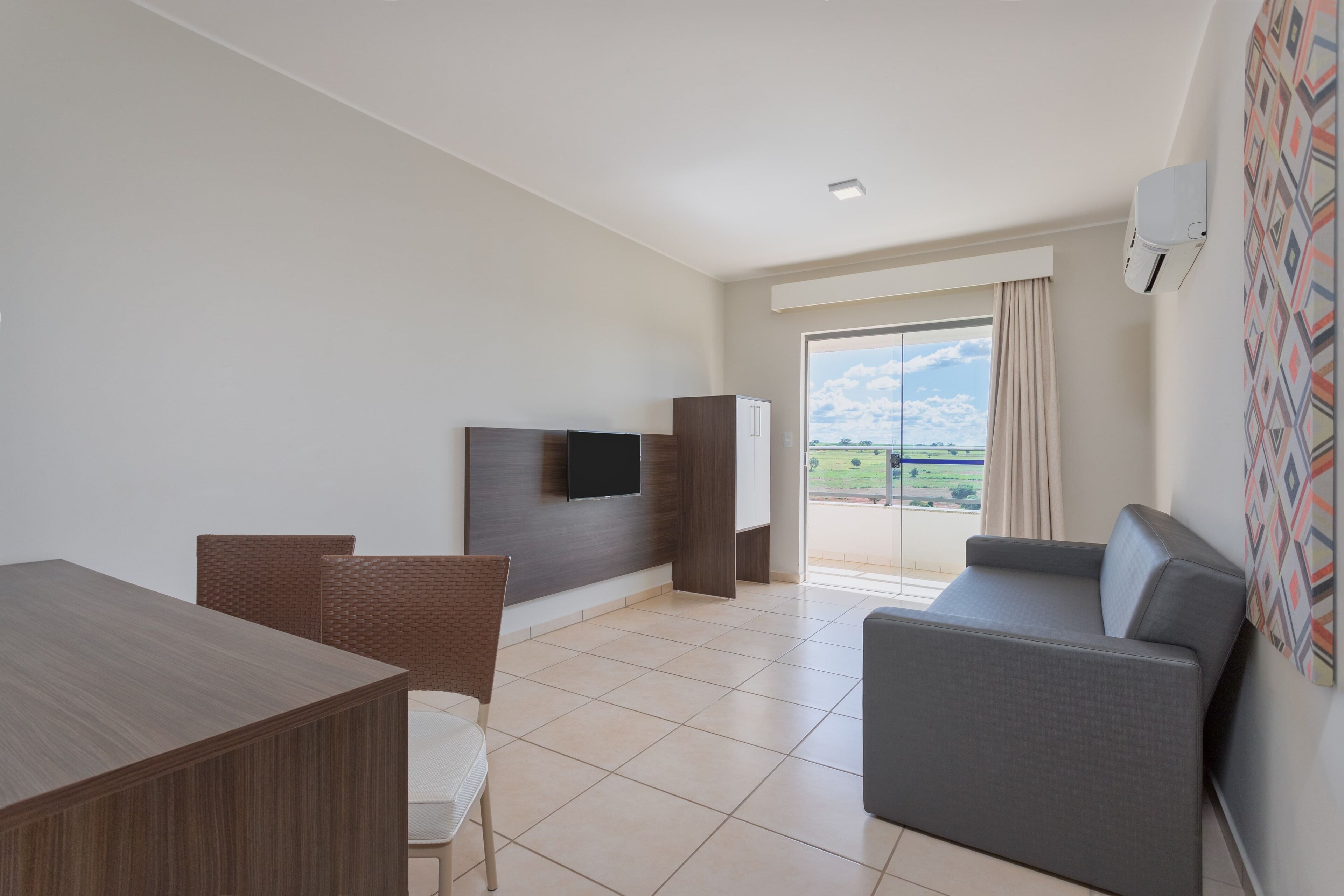 Una habitación de hotel moderna con comedor, sofá, televisor montado en la pared y un amplio ventanal con vistas al exterior.