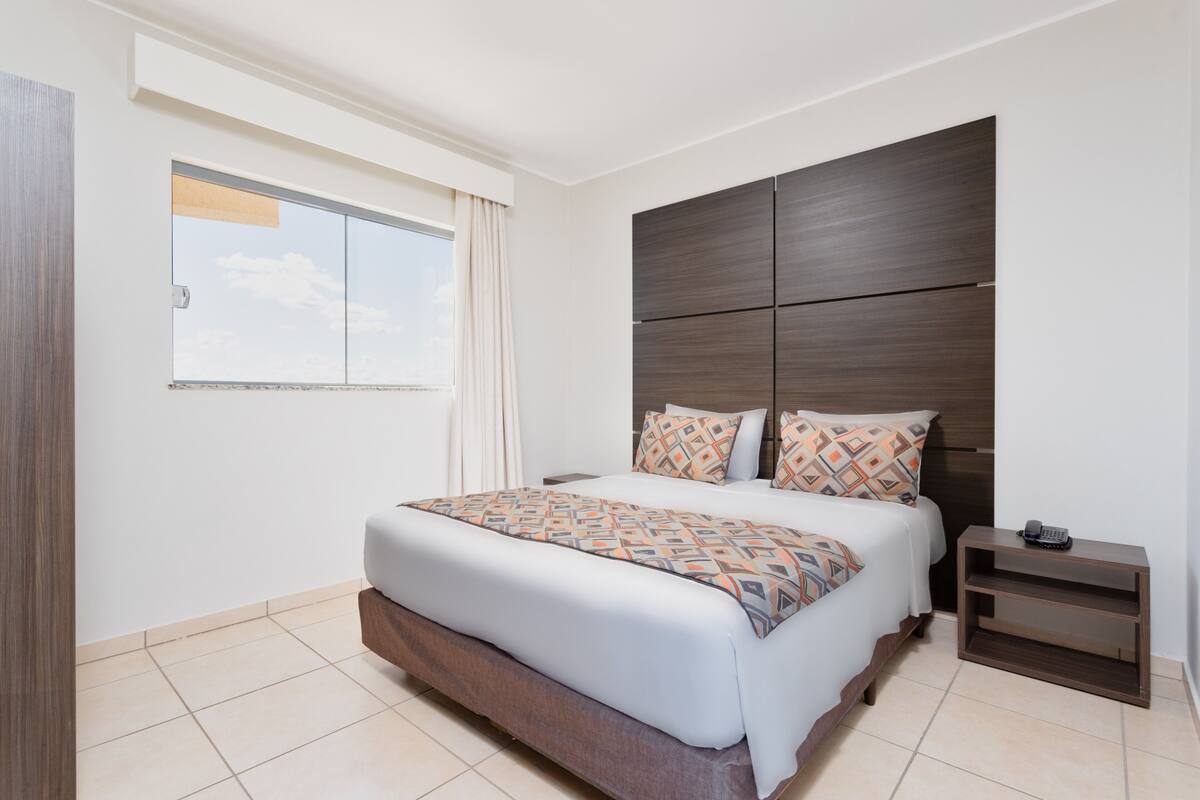 Ein modernes Hotelzimmer mit einem großen Bett, einem dunklen Holz-Kopfbrett, einem kleinen Nachttisch und einem Fenster mit Vorhängen.