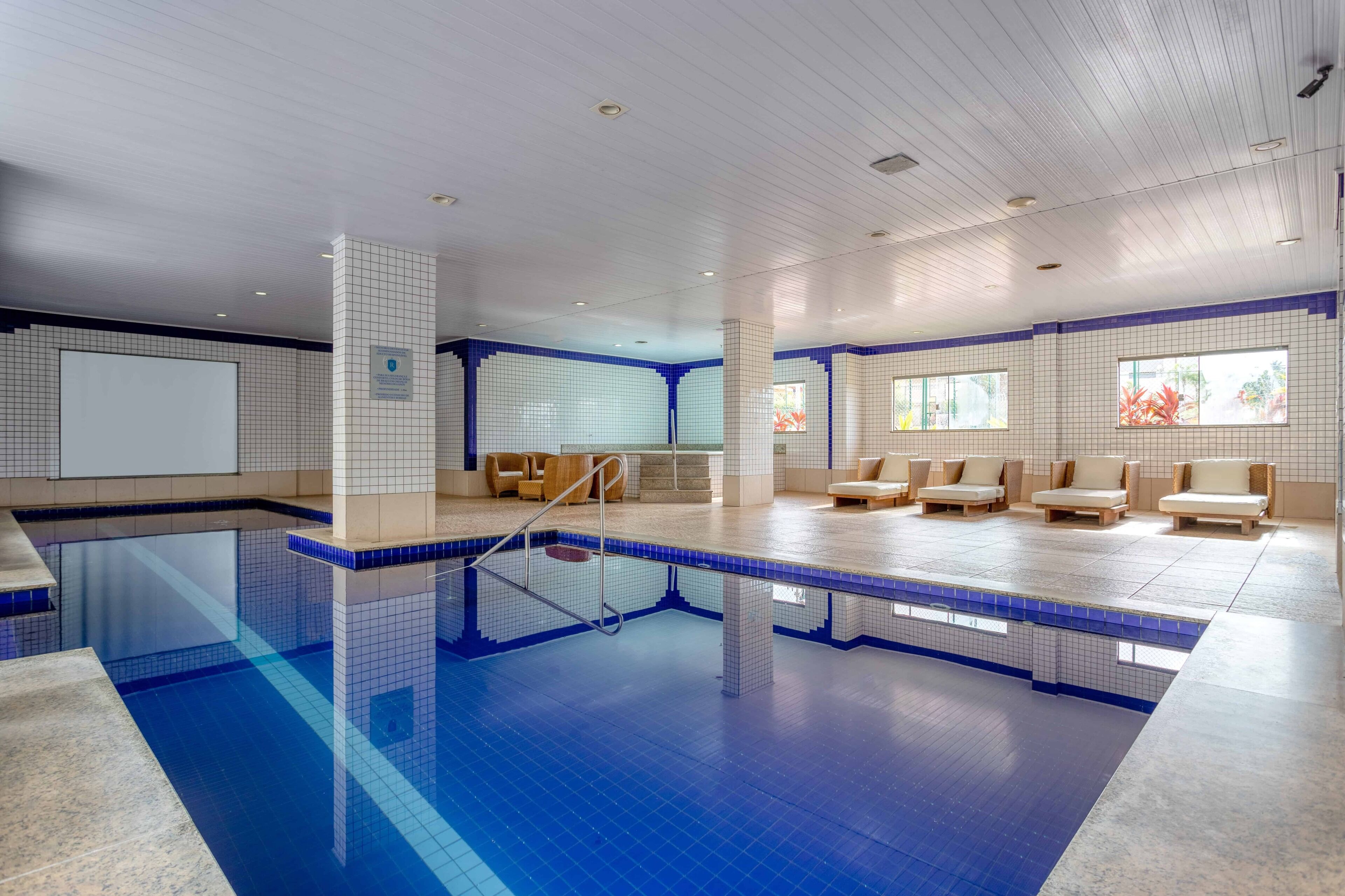 A modern indoor swimming pool with clear blue water, a tiled floor, and a seating area with cushioned chairs.