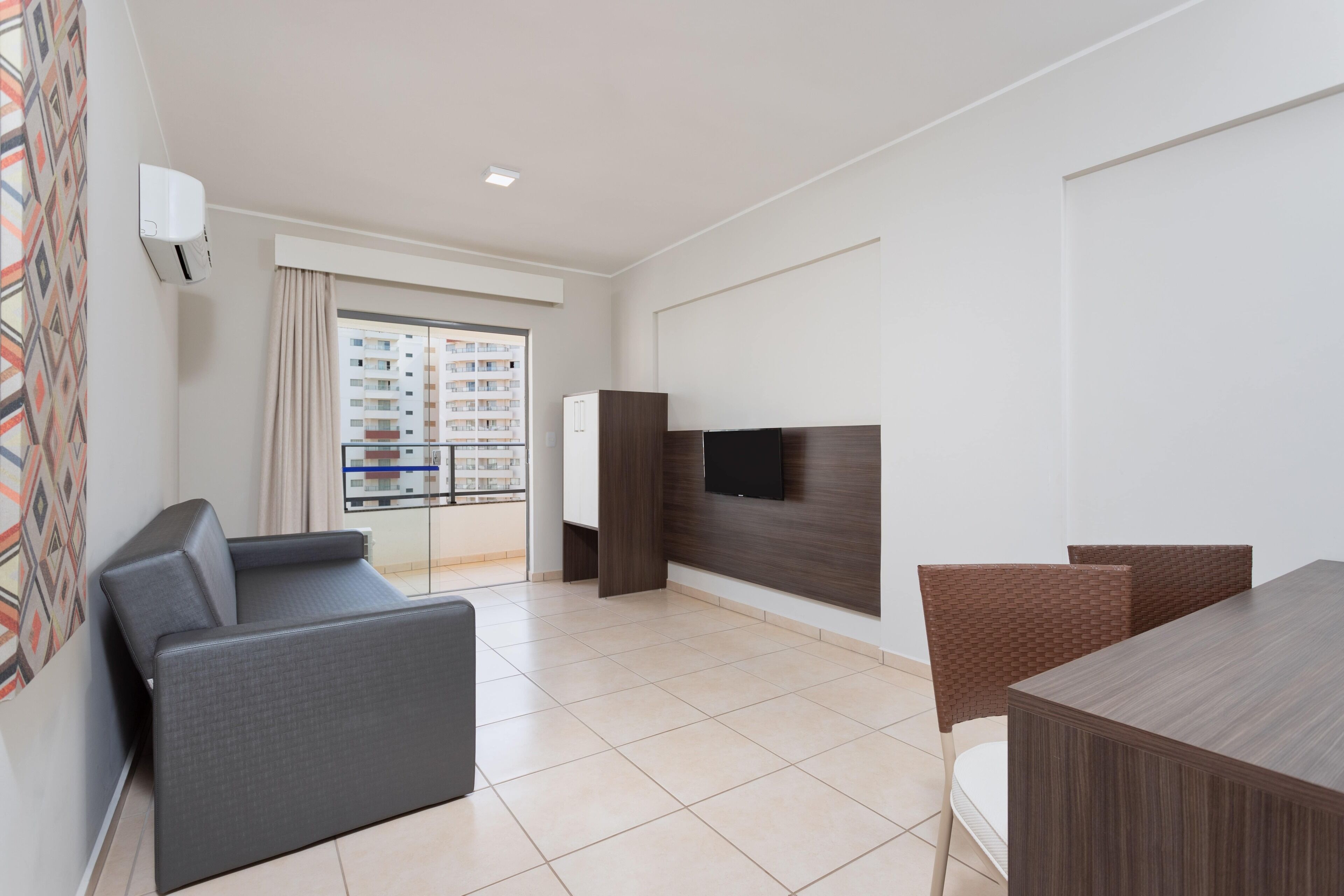 A modern living room with a sofa, a TV mounted on a wooden wall, and a balcony with a view of high-rise buildings.