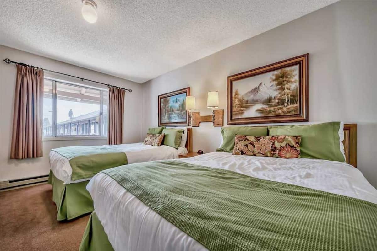 Habitación de hotel con dos camas, ropa de cama verde, un ventanal con cortinas y dos cuadros en la pared.