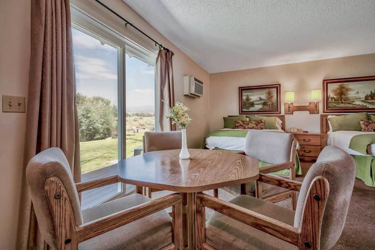 Habitación de hotel con una mesa de comedor, dos camas, una ventana con cortinas y un cuadro en la pared.