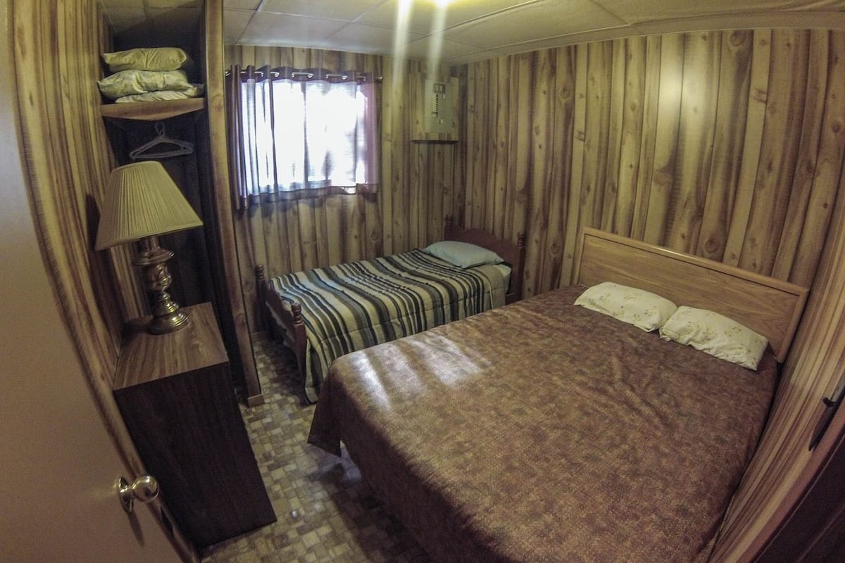 Ein kleines Zimmer mit einem Einzelbett, einem Nachttisch mit Lampe, einem kleinen Kleiderschrank und einem Fenster mit Vorhängen.