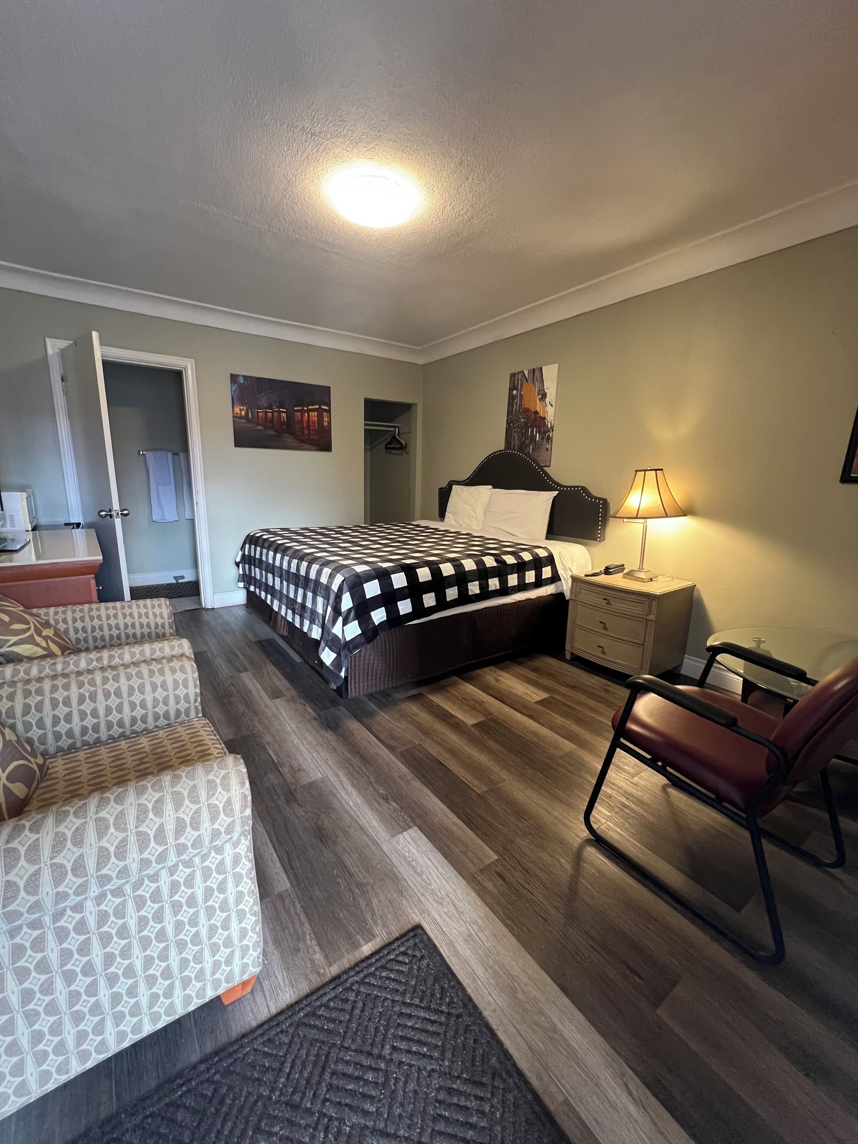 Ein Hotelzimmer mit einem Bett, einem Sessel, einem Nachttisch, einer Lampe, einer Kommode und einer Tür zu einem anderen Raum.