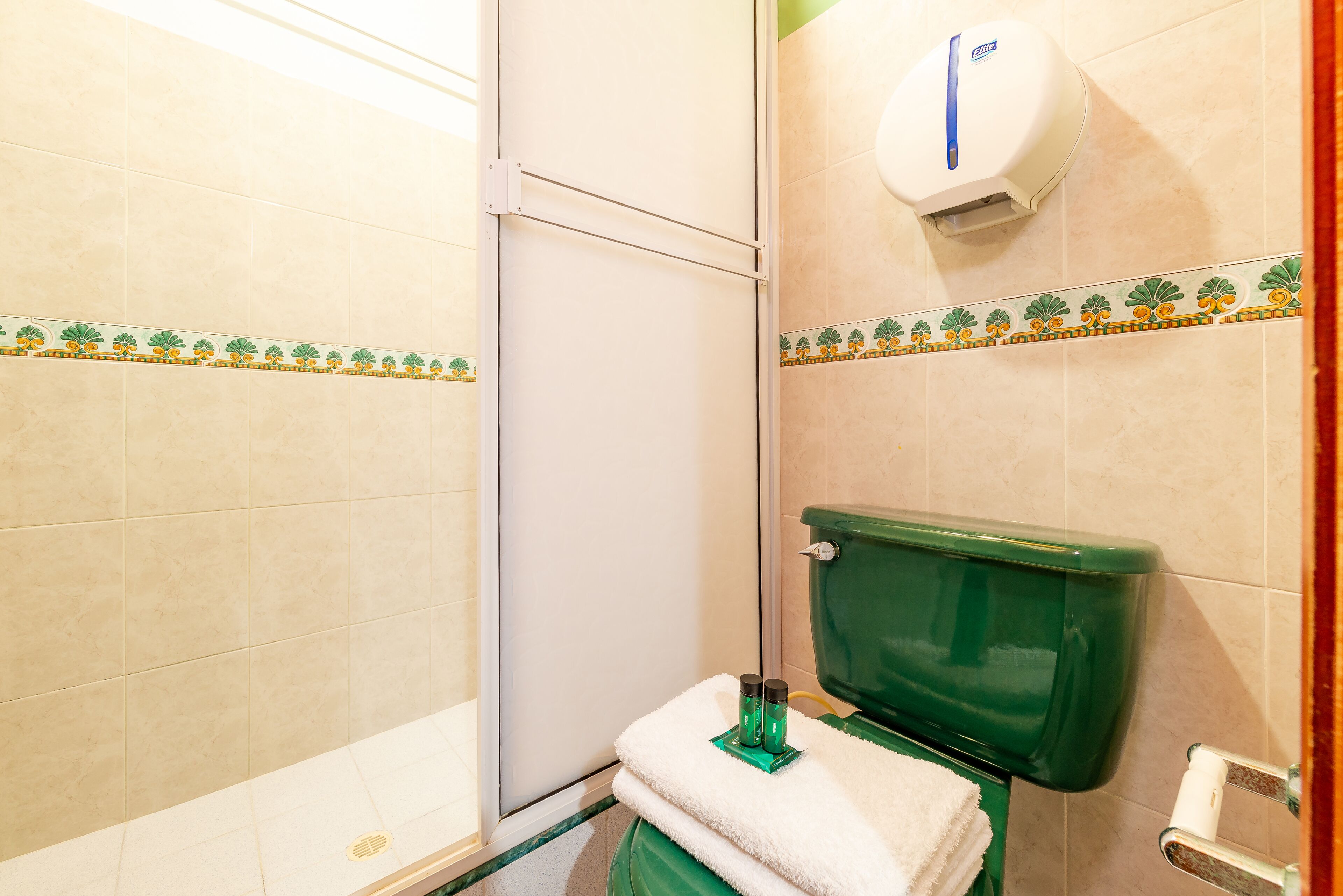 A bathroom with beige tiles, a green toilet, a white towel, and a hand dryer.