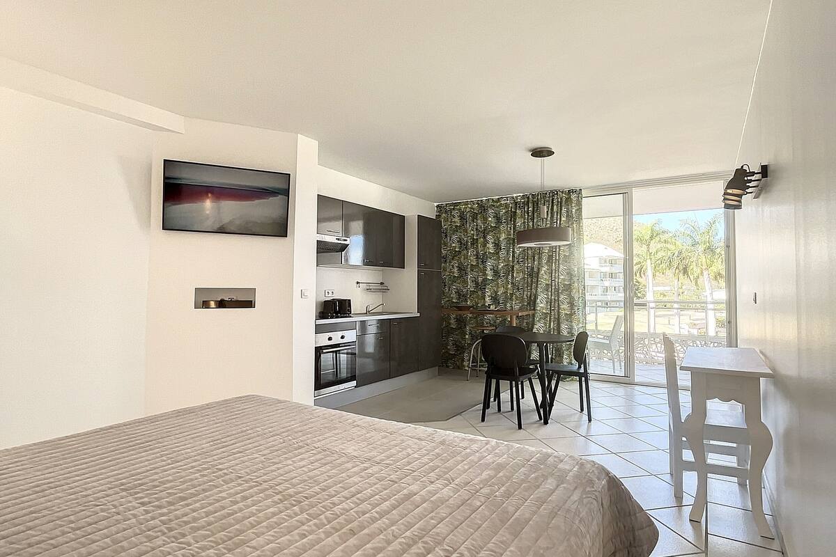 A modern hotel room with a large bed, a kitchenette, a dining area, and a balcony with a view.