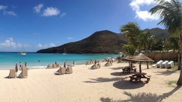 A beach with sun loungers and umbrellas, a wooden deck, and a mountain in the background.