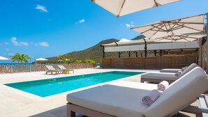 A swimming pool with lounge chairs and umbrellas, surrounded by a wooden fence and a mountain backdrop. - Le Domaine Anse Marcel Beach Resort (Anse Marcel)