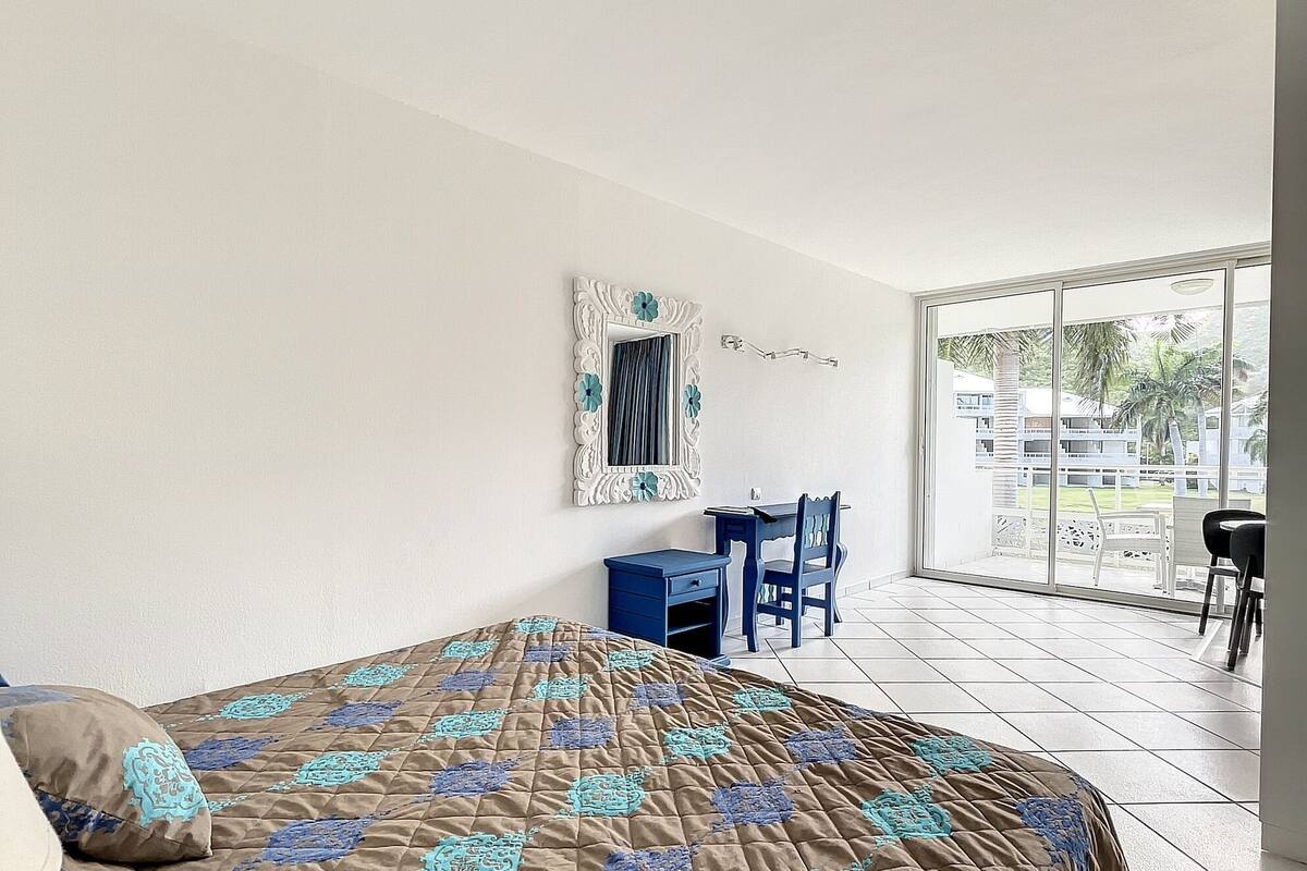 A hotel room with a large bed, a desk, and a chair. There is a window with a view of palm trees and buildings.