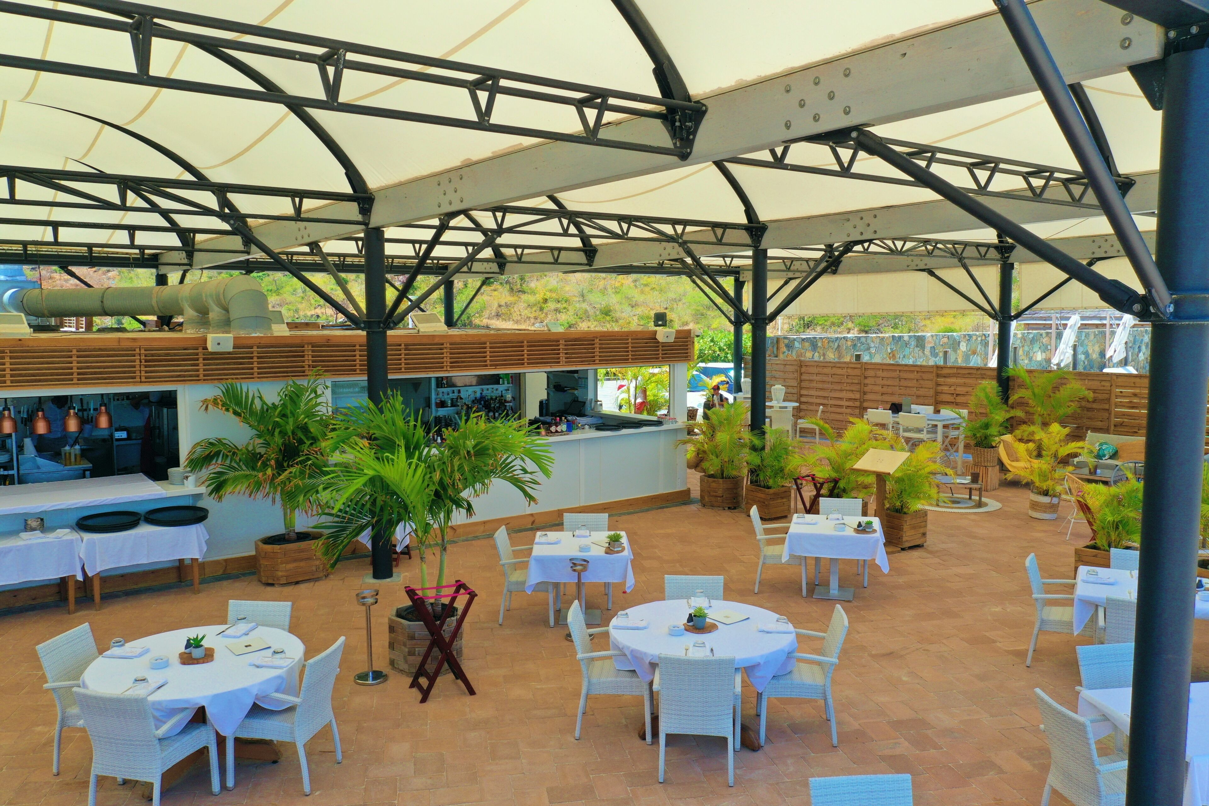 An outdoor patio area with round tables and chairs, a bar area, and potted plants.