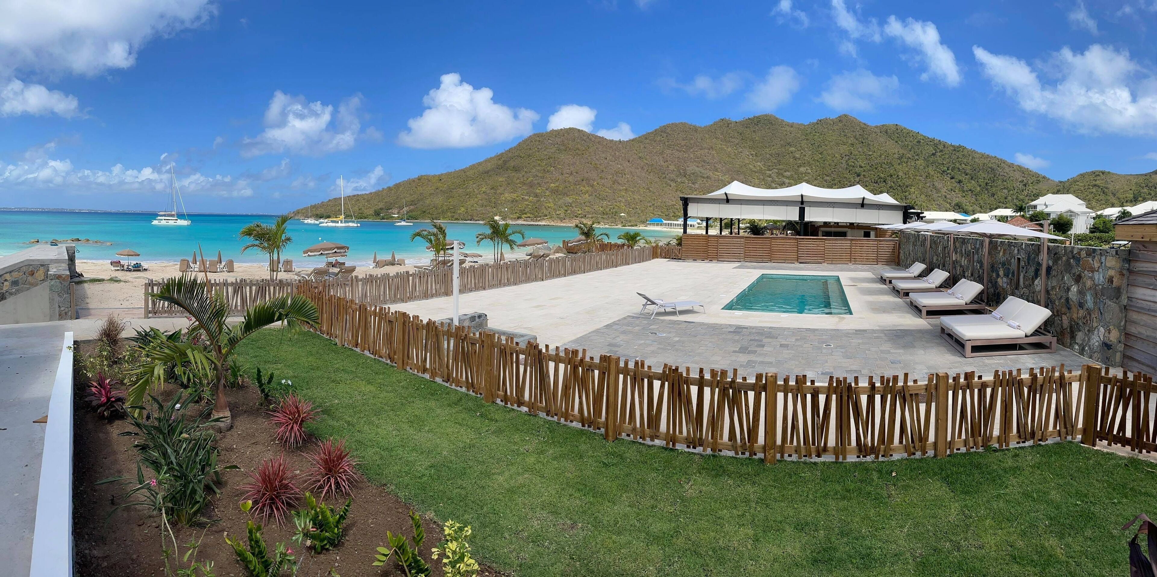 A pool area with lounge chairs, a wooden fence, and a view of the beach and mountains.