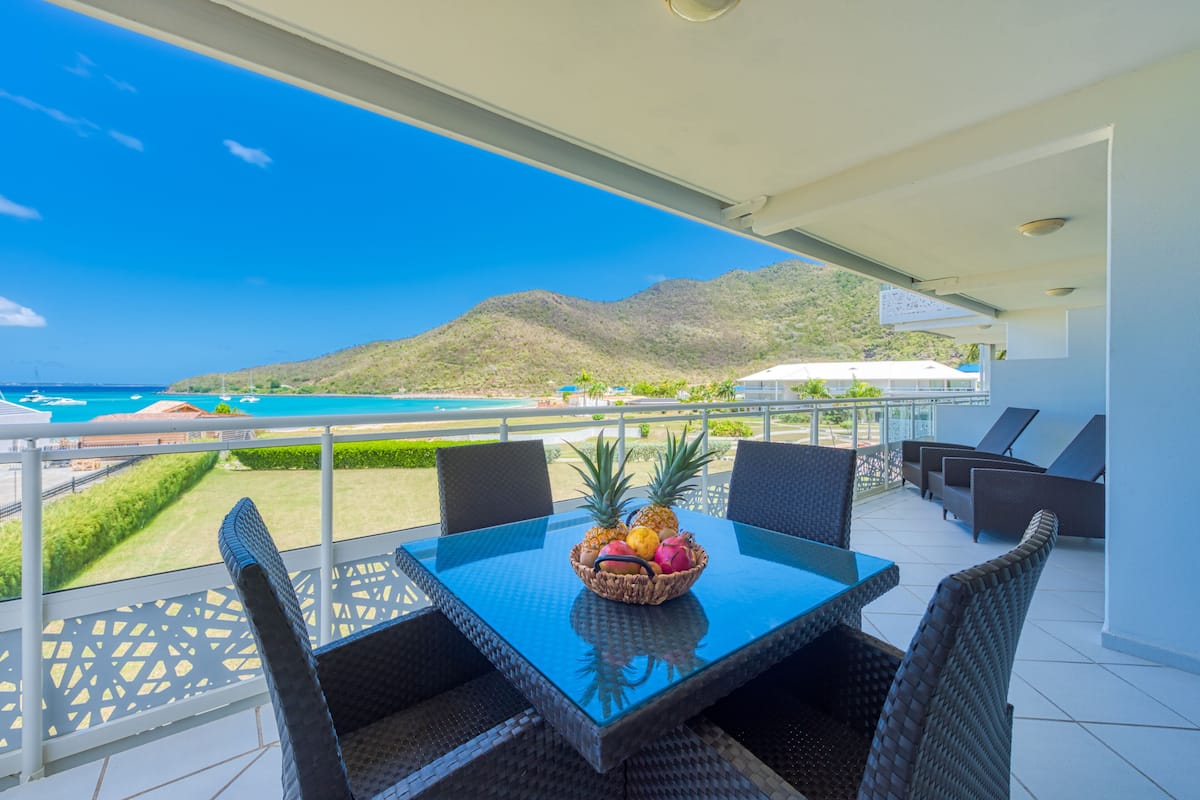 A balcony with a table and chairs, a fruit basket, and a view of the ocean and mountains.