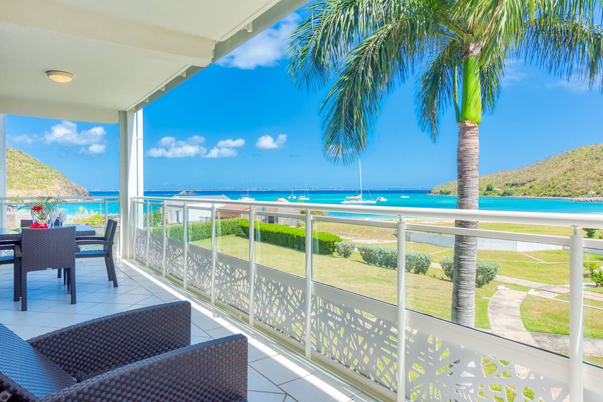 A balcony with a view of the ocean, a palm tree, and a dining area with chairs and a table.