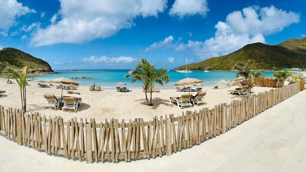 A beach with sun loungers and umbrellas, a wooden fence, and a view of the sea and hills. - Le Domaine Anse Marcel Beach Resort (Anse Marcel)