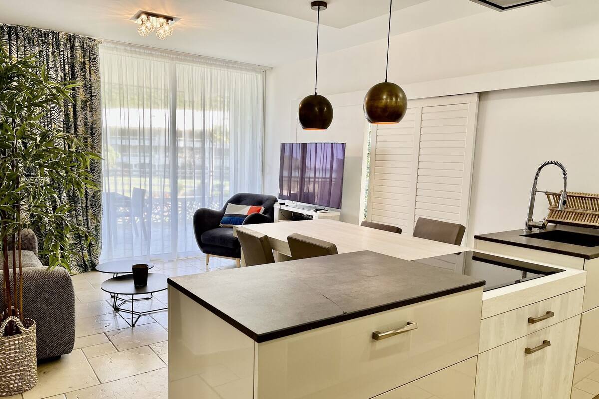A modern kitchen with a dining area, large windows, and a potted plant.
