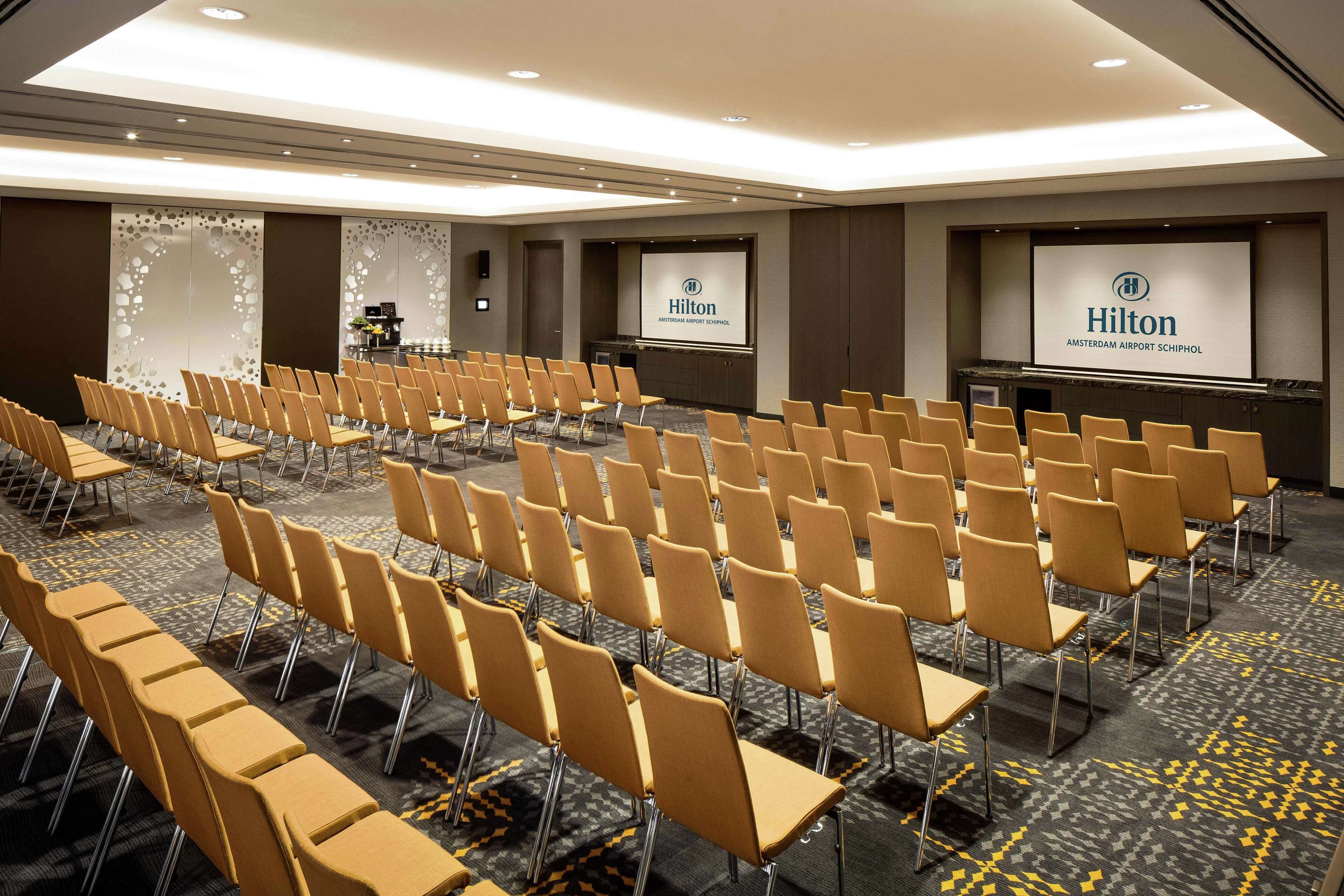 A conference room with rows of chairs, a podium, and a screen displaying the Hilton logo.