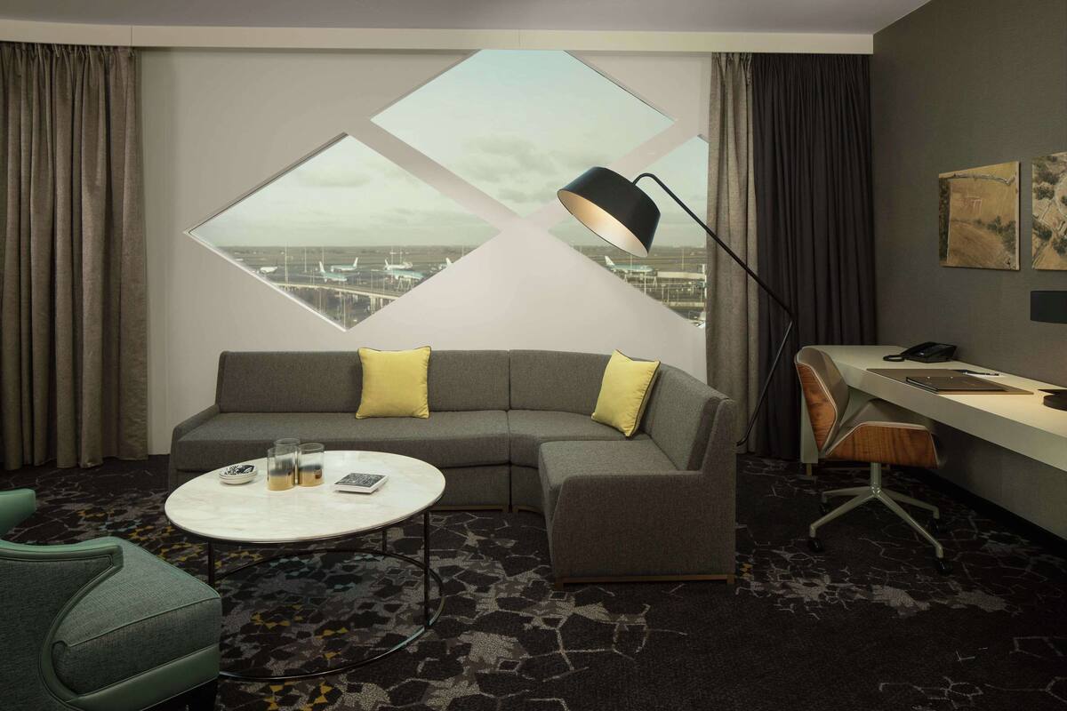 Una habitación de hotel moderna con sofá modular, una mesa de centro redonda, un escritorio con una silla y un ventanal con vistas al aeropuerto.