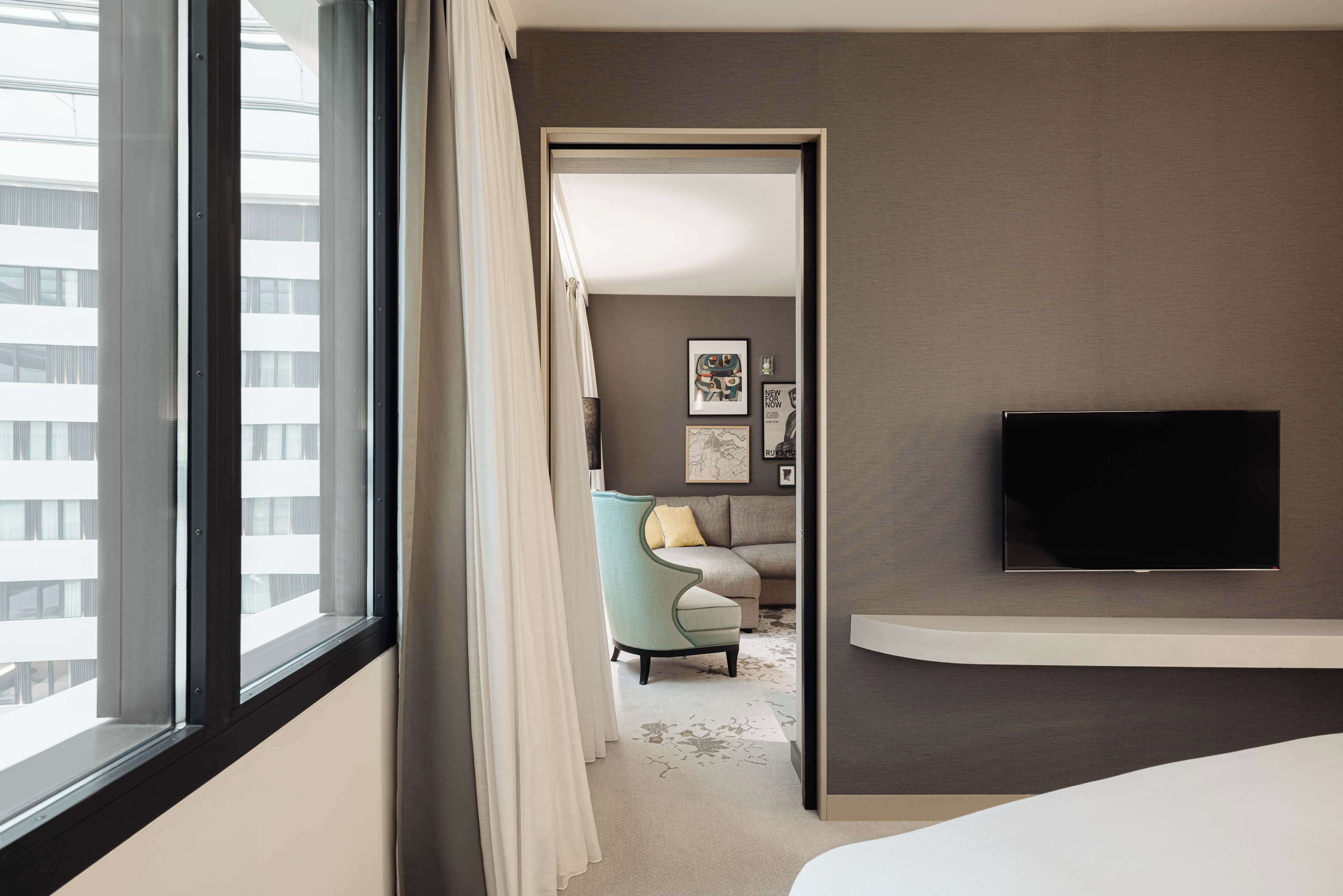 A hotel room with a large window, a flat-screen TV mounted on the wall, a sofa, and a chair.