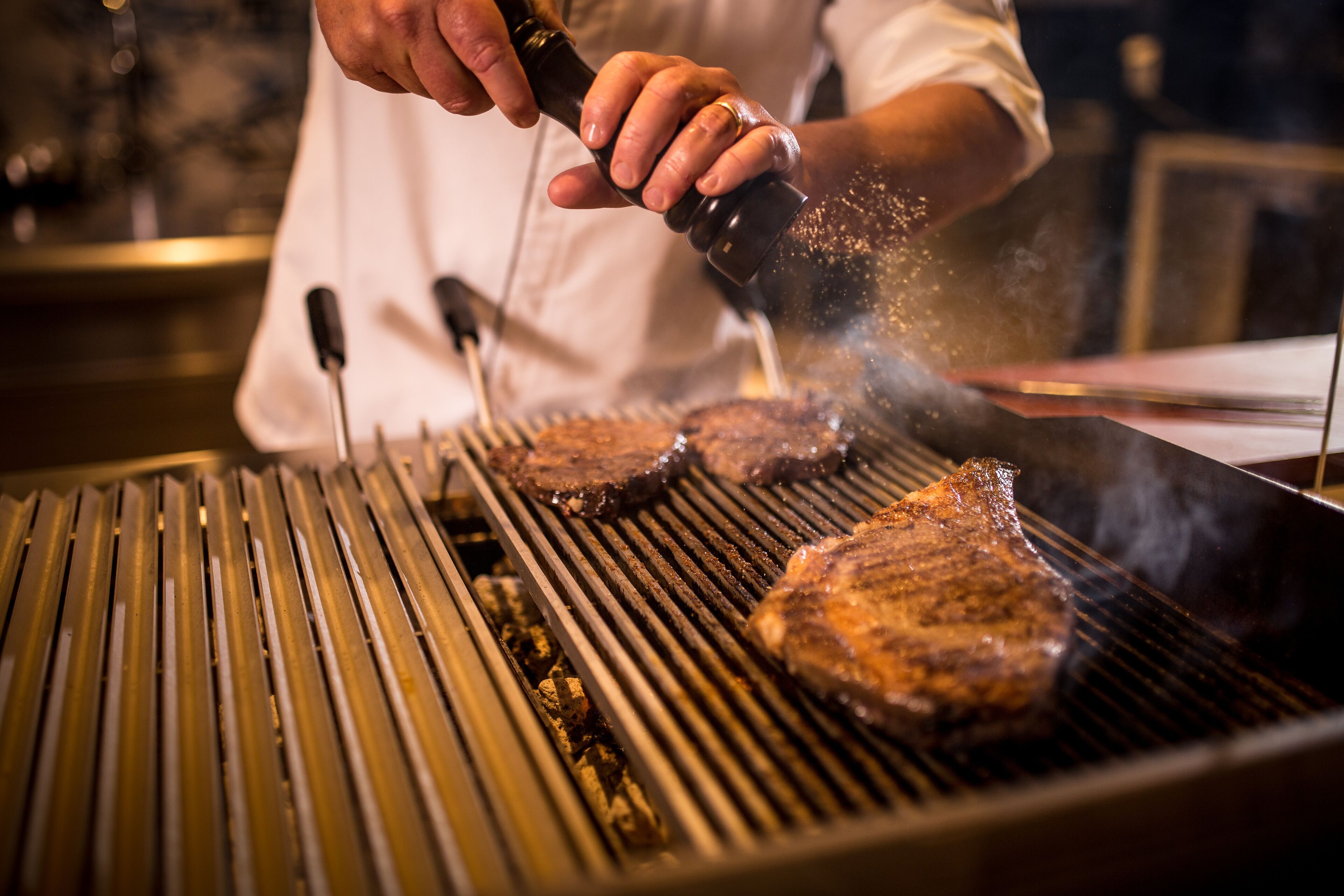 Una persona asando filetes a la parrilla, con marcas visibles de la parrilla y humo.