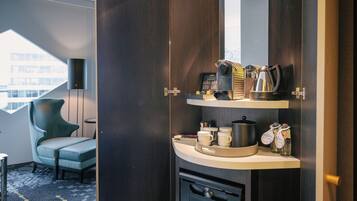 Habitación de hotel con un armario de madera oscura, una kitchenette pequeña con cafetera, tazas y condimentos. Hay una silla verde y vista a un edificio a través de una ventana.