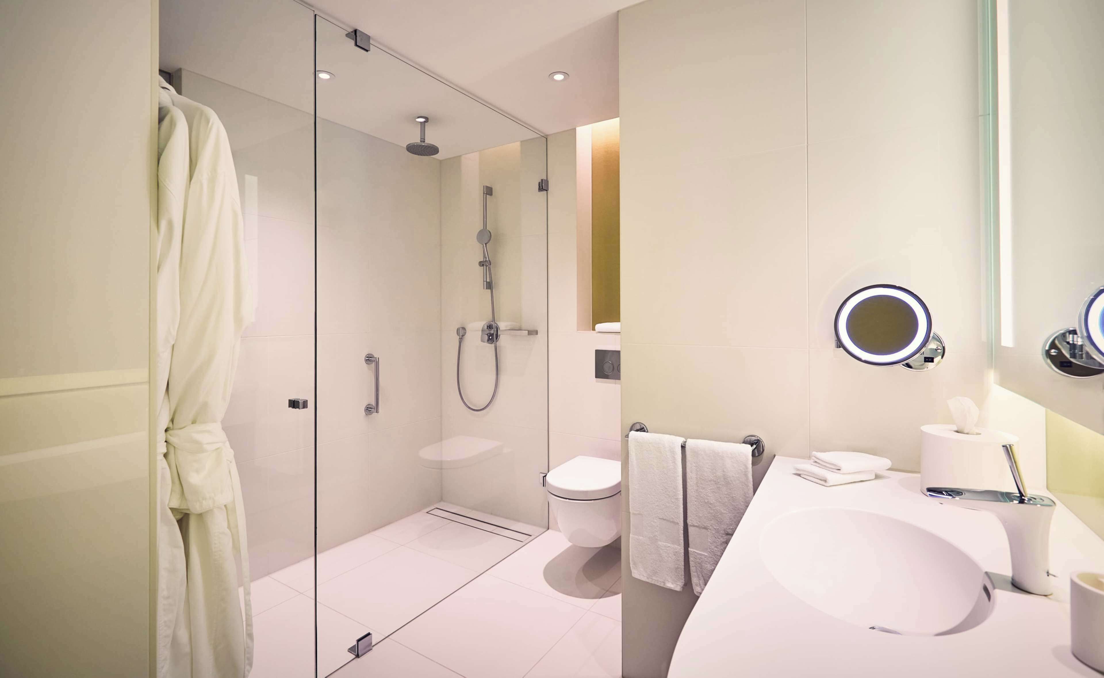 Un baño moderno con ducha de vidrio, lavamanos blanco, inodoro y espejo con luz de aro.