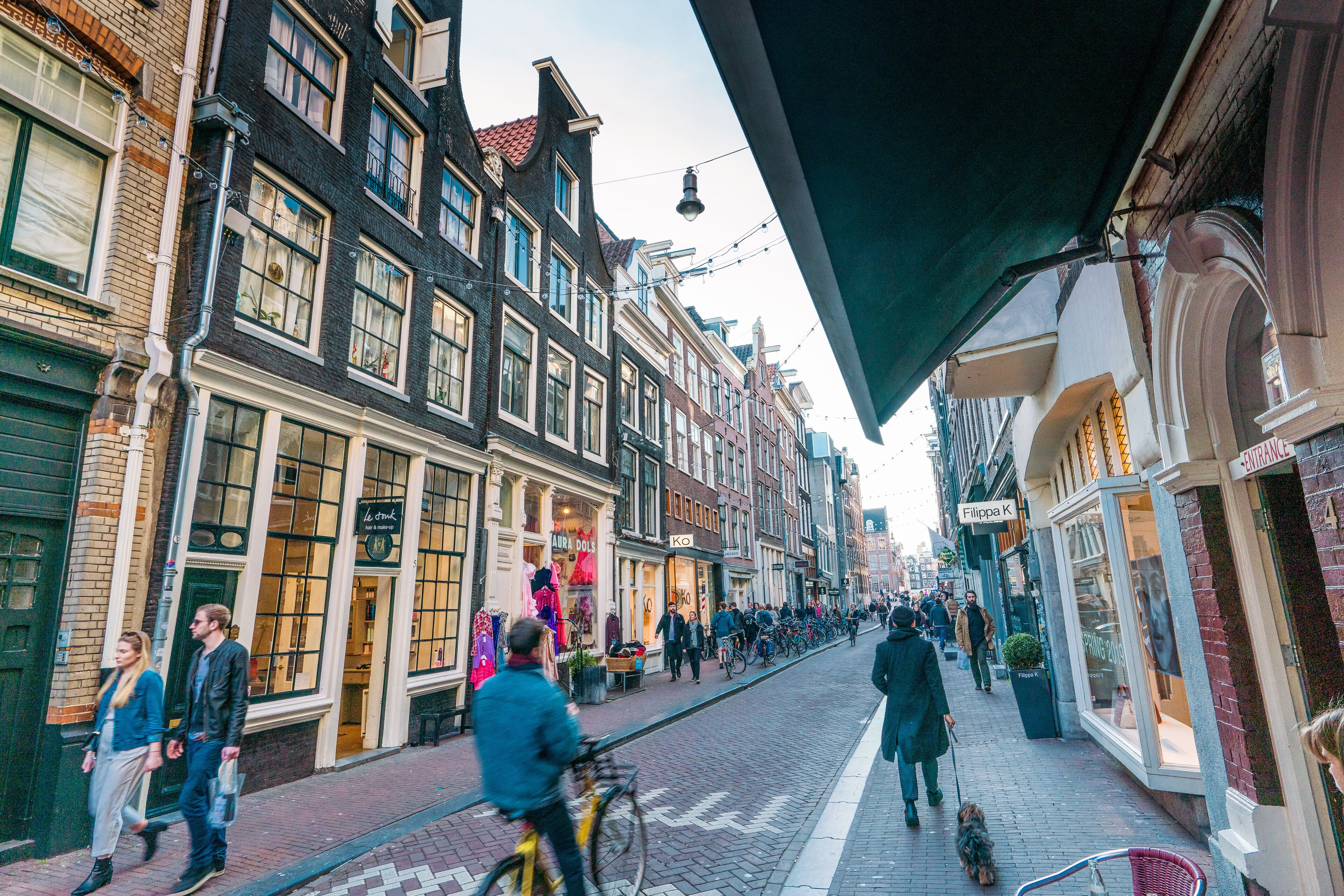 A street lined with historic buildings, some with shops and signs, and people walking and cycling.