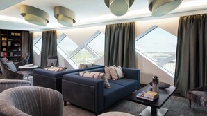 Un salon moderne comprenant un ensemble de canapés, une table basse et une bibliothèque. De grandes fenêtres, dotées de rideaux, laissent entrer la lumière naturelle et offrent une vue sur la ville.
