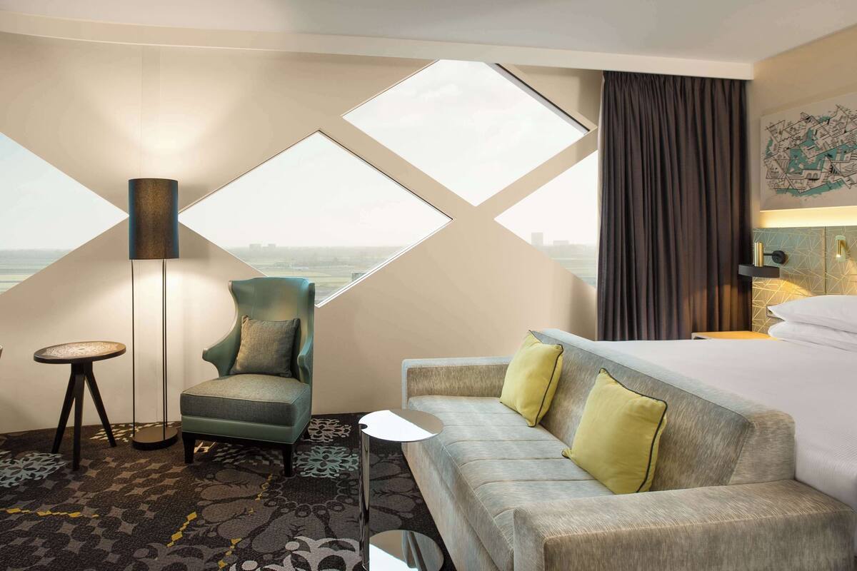 Habitación de hotel con un ventanal grande, un sofá, una silla, un escritorio y una lámpara.