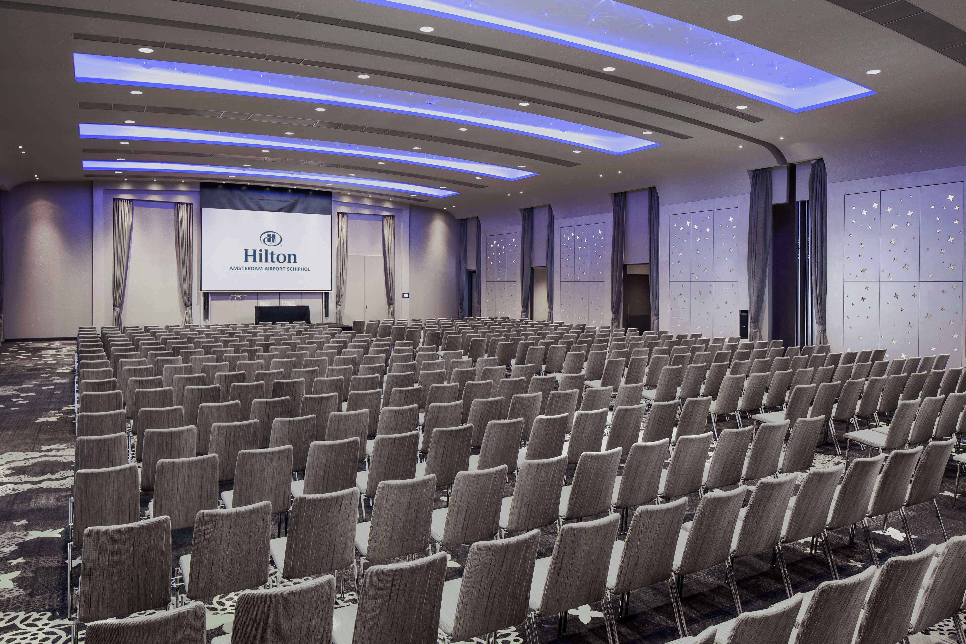 A large, modern conference room with rows of chairs, a stage with a Hilton sign, and a star-patterned wall.