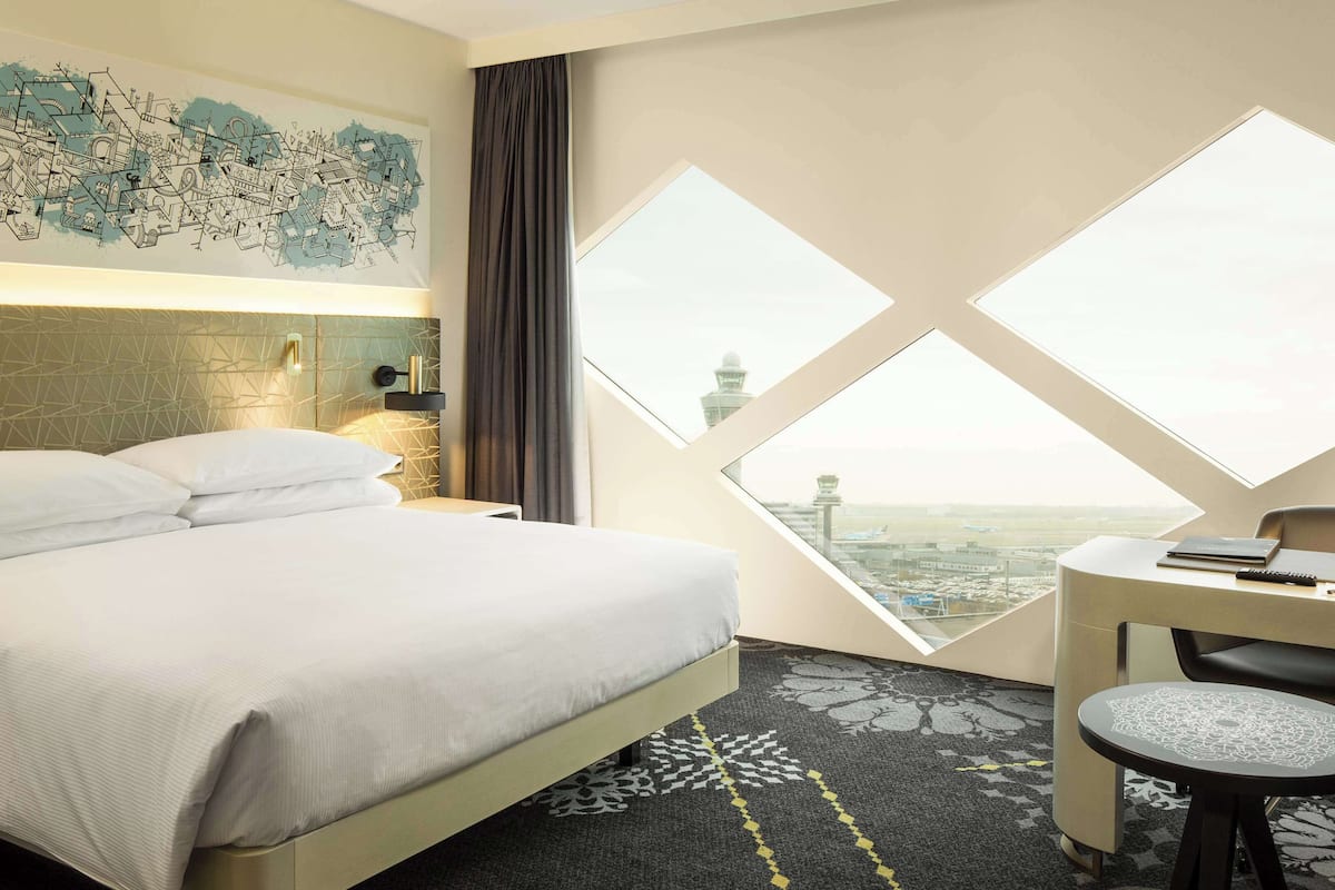 Habitación de hotel con cama, escritorio y silla, y un ventanal que ofrece vistas al aeropuerto.