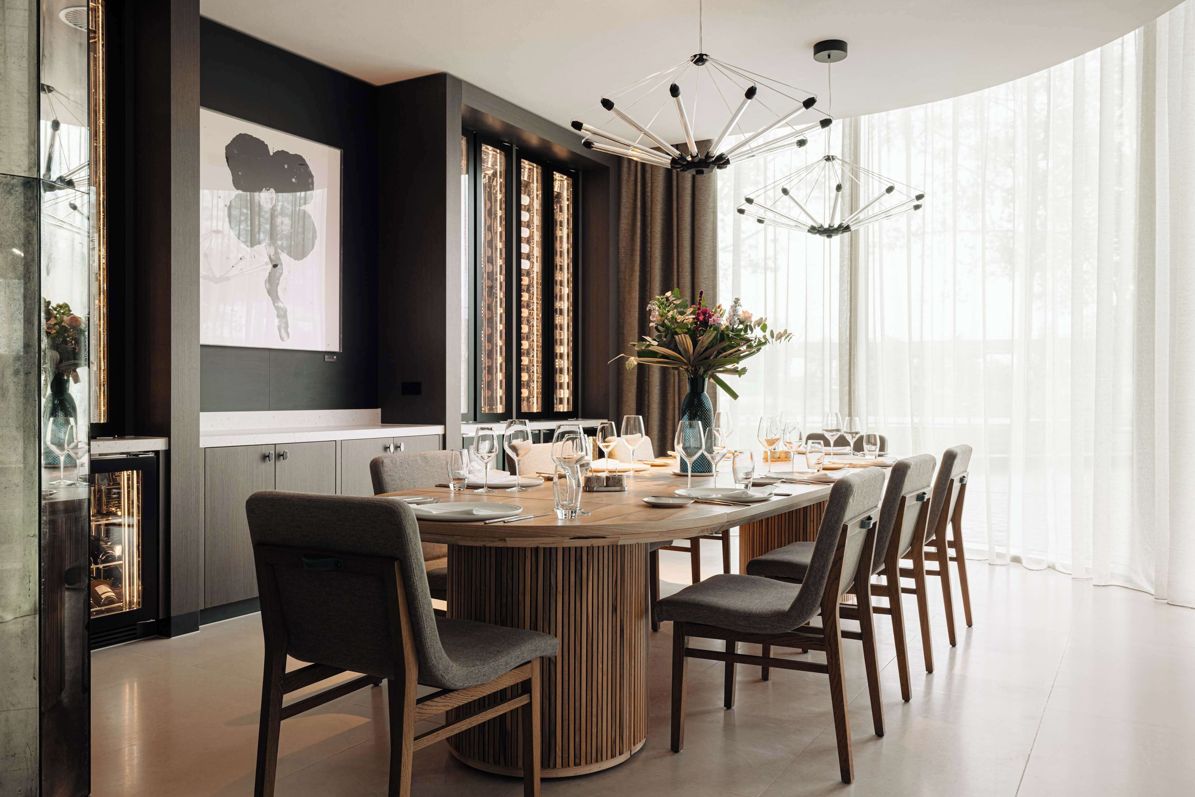 Uma área de jantar moderna com mesa redonda de madeira, cadeiras escuras e um lustre contemporâneo.