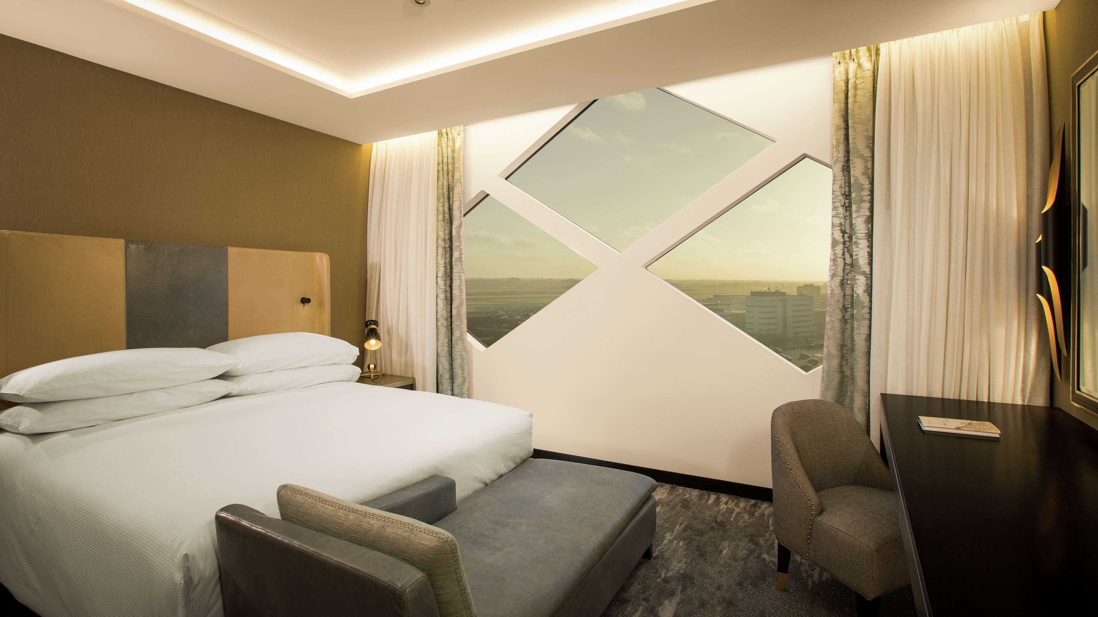 Habitación de hotel con cama, dos sillones, escritorio y ventana grande con vista a la ciudad.