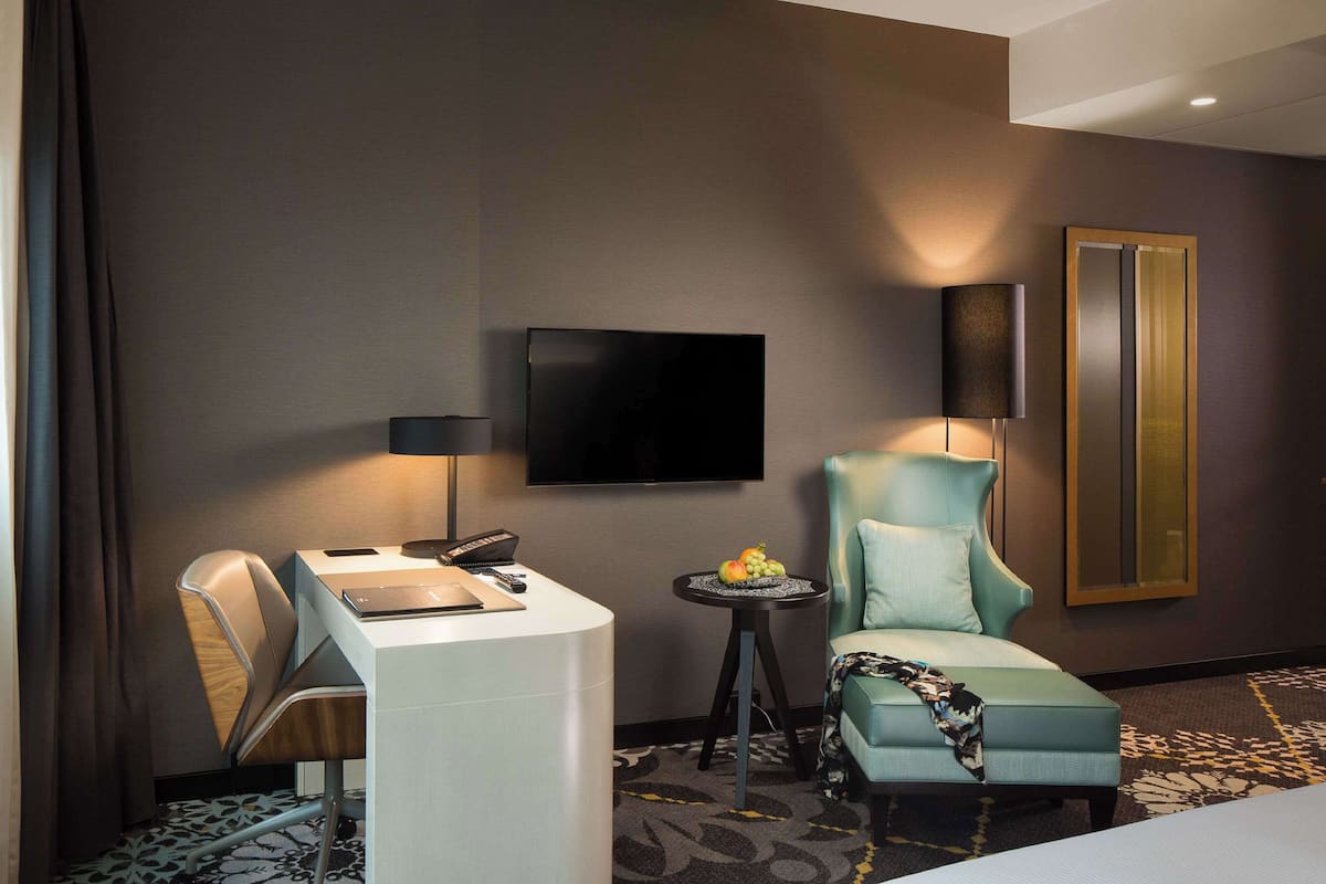 Una habitación de hotel moderna con una televisión de pantalla plana en la pared, un sillón cómodo, un escritorio con silla y una mesita de noche con fruta.