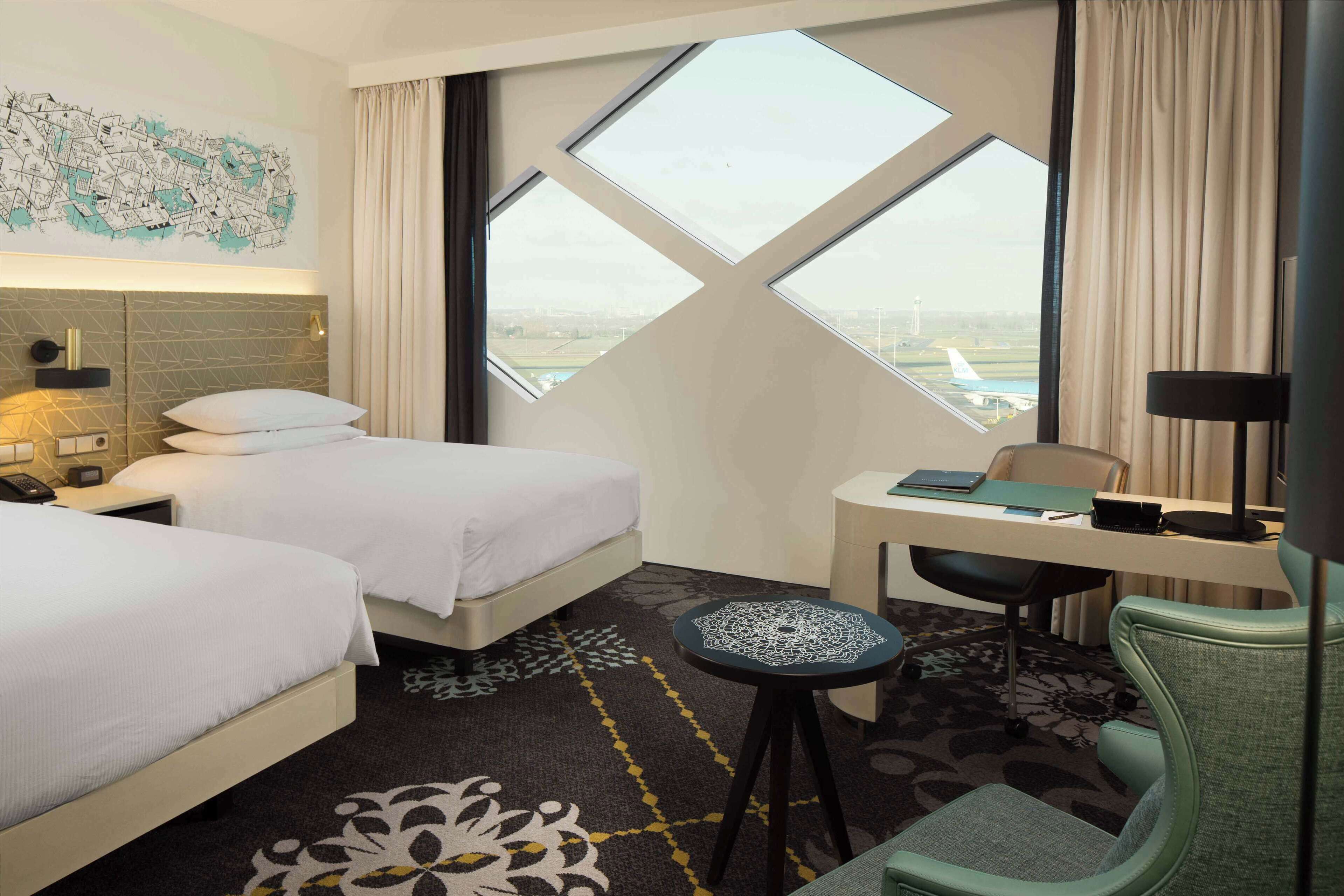 Ett hotellrum med två sängar, ett skrivbord, en stol, en lampa och ett stort fönster med utsikt över en flygplats.