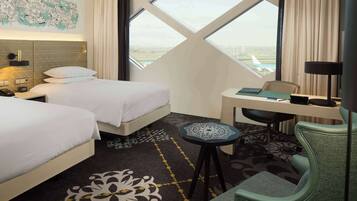 Quarto de hotel com duas camas, uma escrivaninha, uma cadeira, um abajur e uma janela grande com vista para um aeroporto.