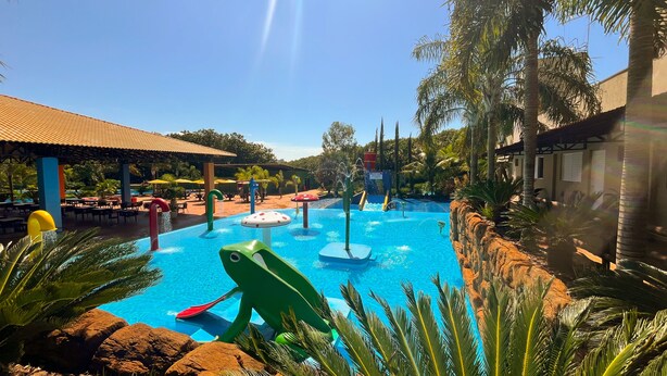A swimming pool with water features, surrounded by palm trees and a covered area with tables and umbrellas. - Hotel Dolce Dulce (Olímpia)