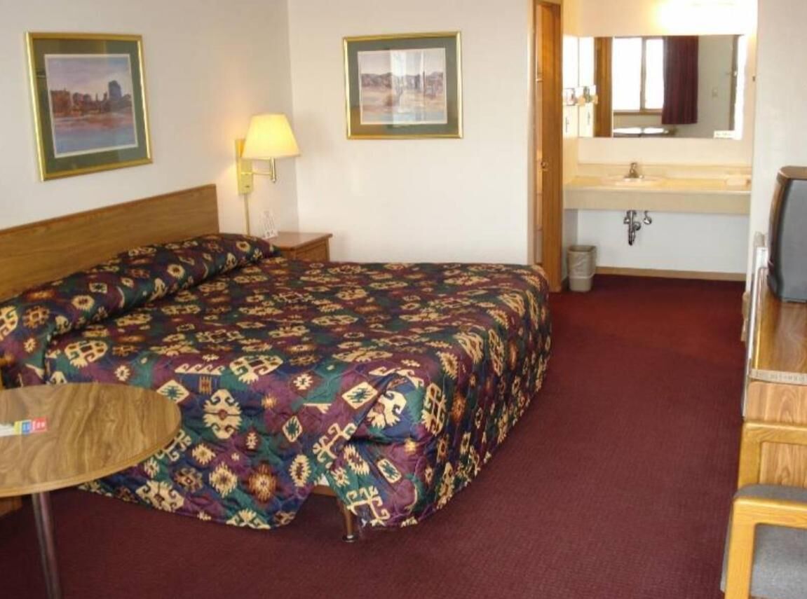 A hotel room with a large bed, a bedside table with a lamp, a small round table, a mirror above the sink, and a view of a cityscape.