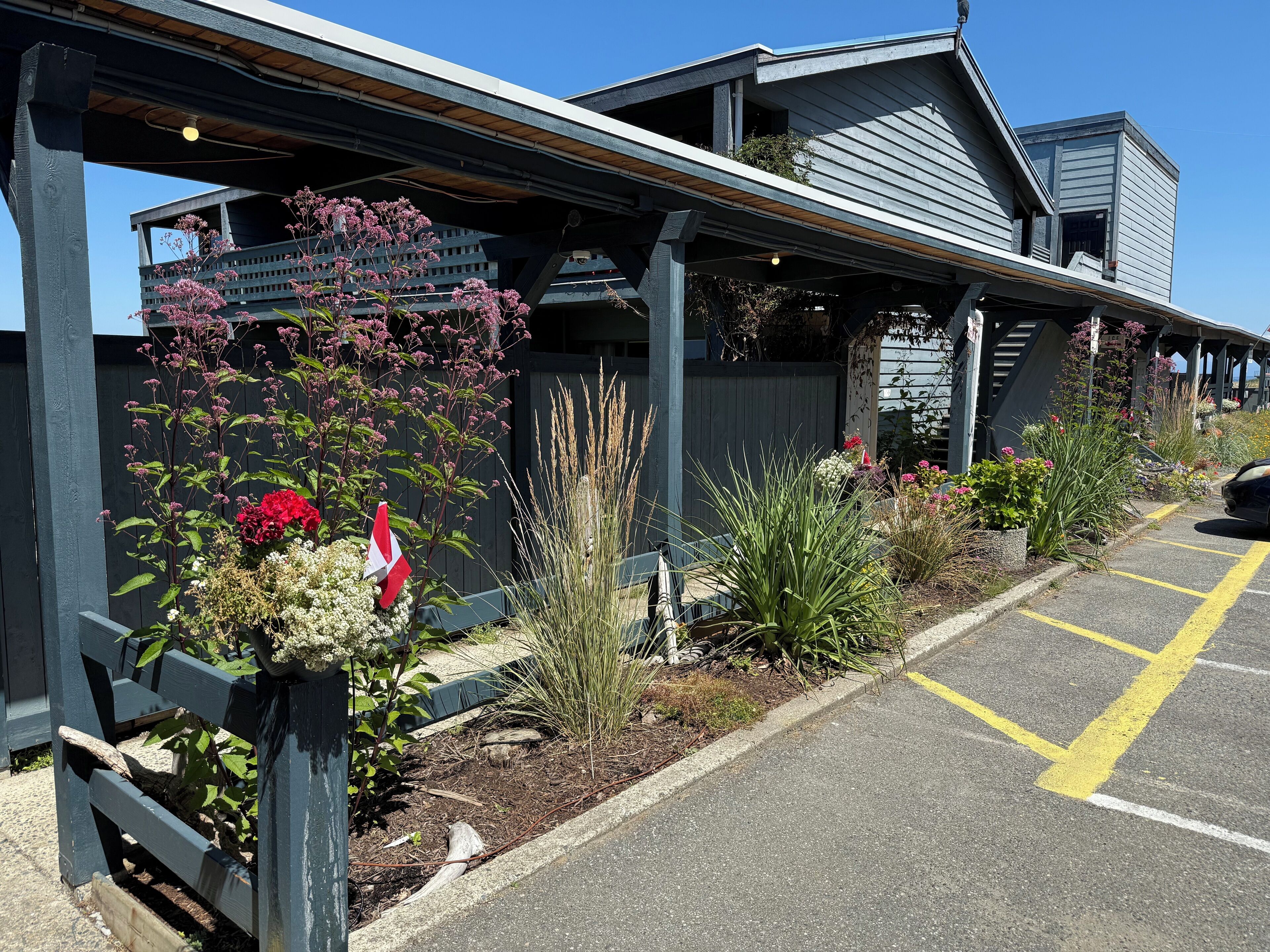 A building with a covered walkway, a parking area, and various plants and flowers.
