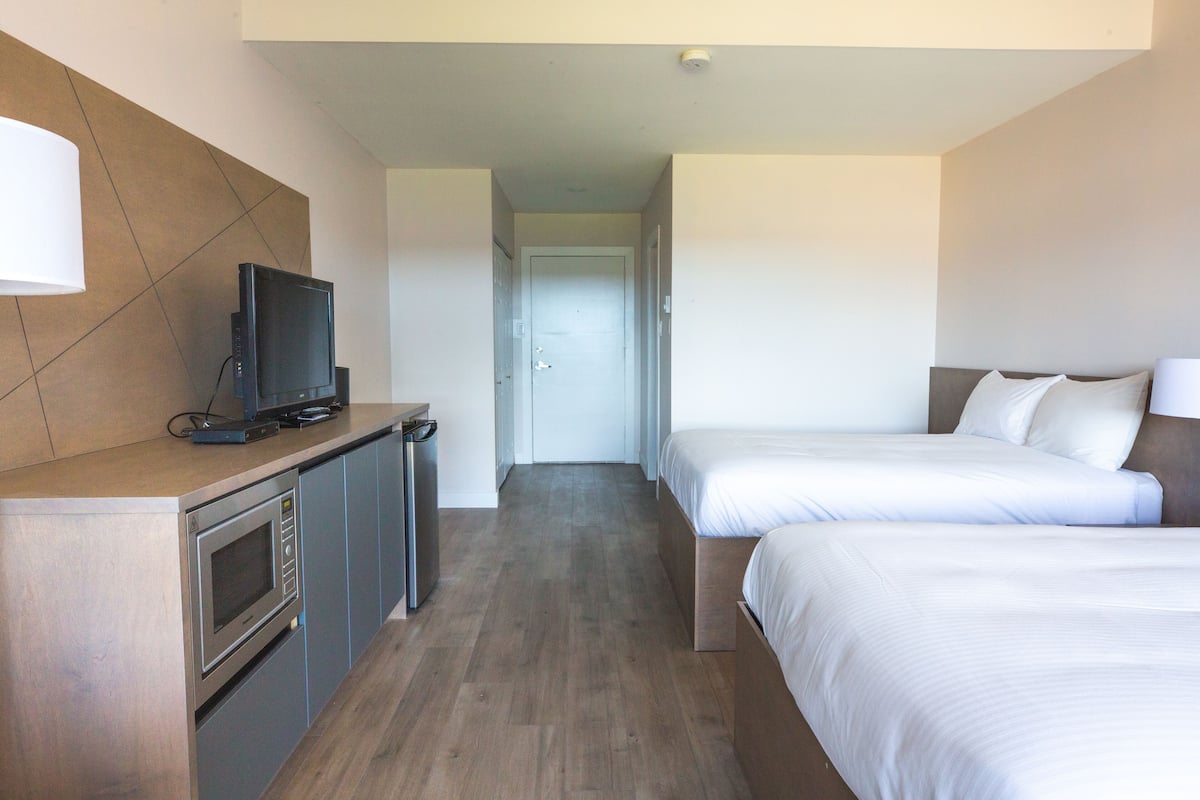 A hotel room with a kitchenette, a TV, two beds, and a door leading to a hallway.