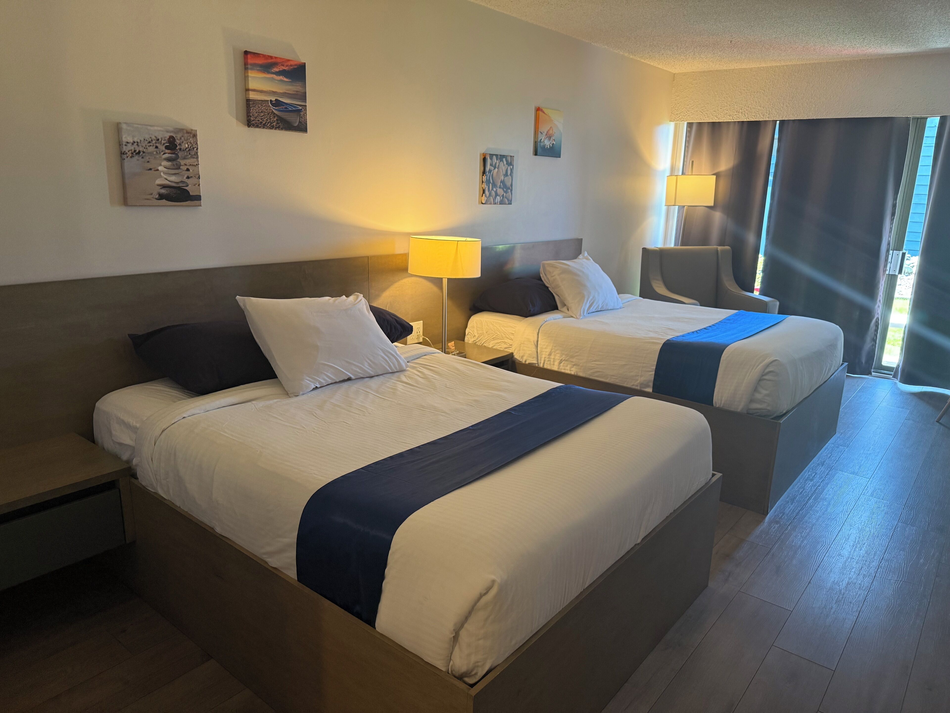 A hotel room with two beds, a nightstand, a lamp, and artwork on the walls.