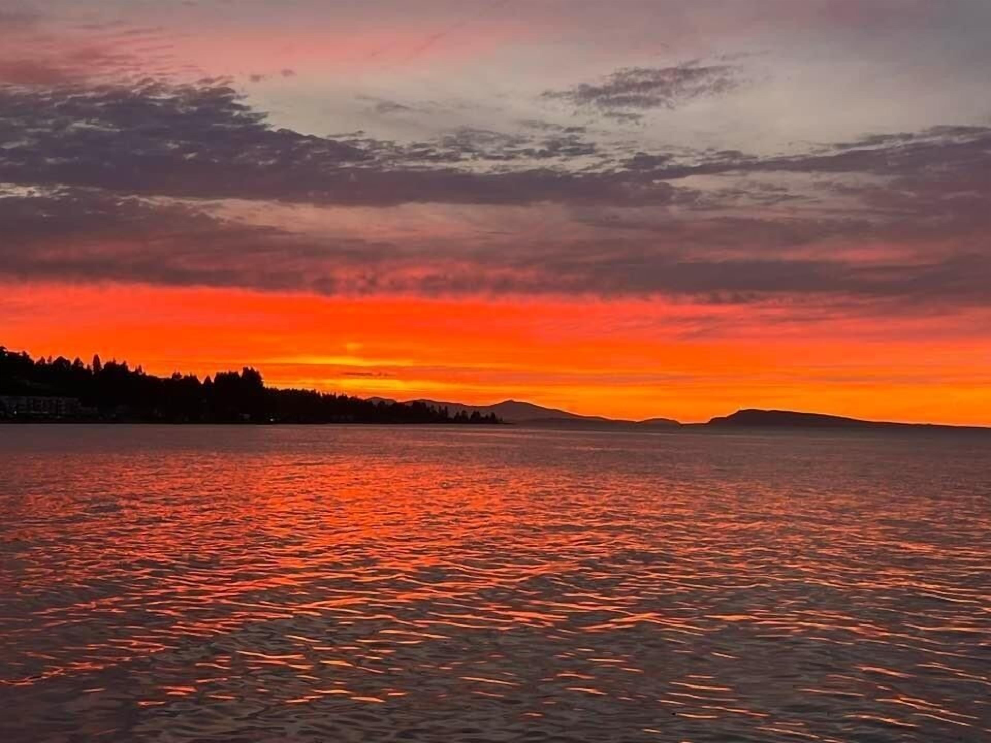 A sunset over a calm body of water with a silhouette of a forested coastline and distant hills.