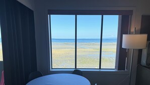 Une pièce dotée d’une grande fenêtre donnant sur une plage. On y trouve une table ronde entourée de chaises, une lampe et un radiateur.