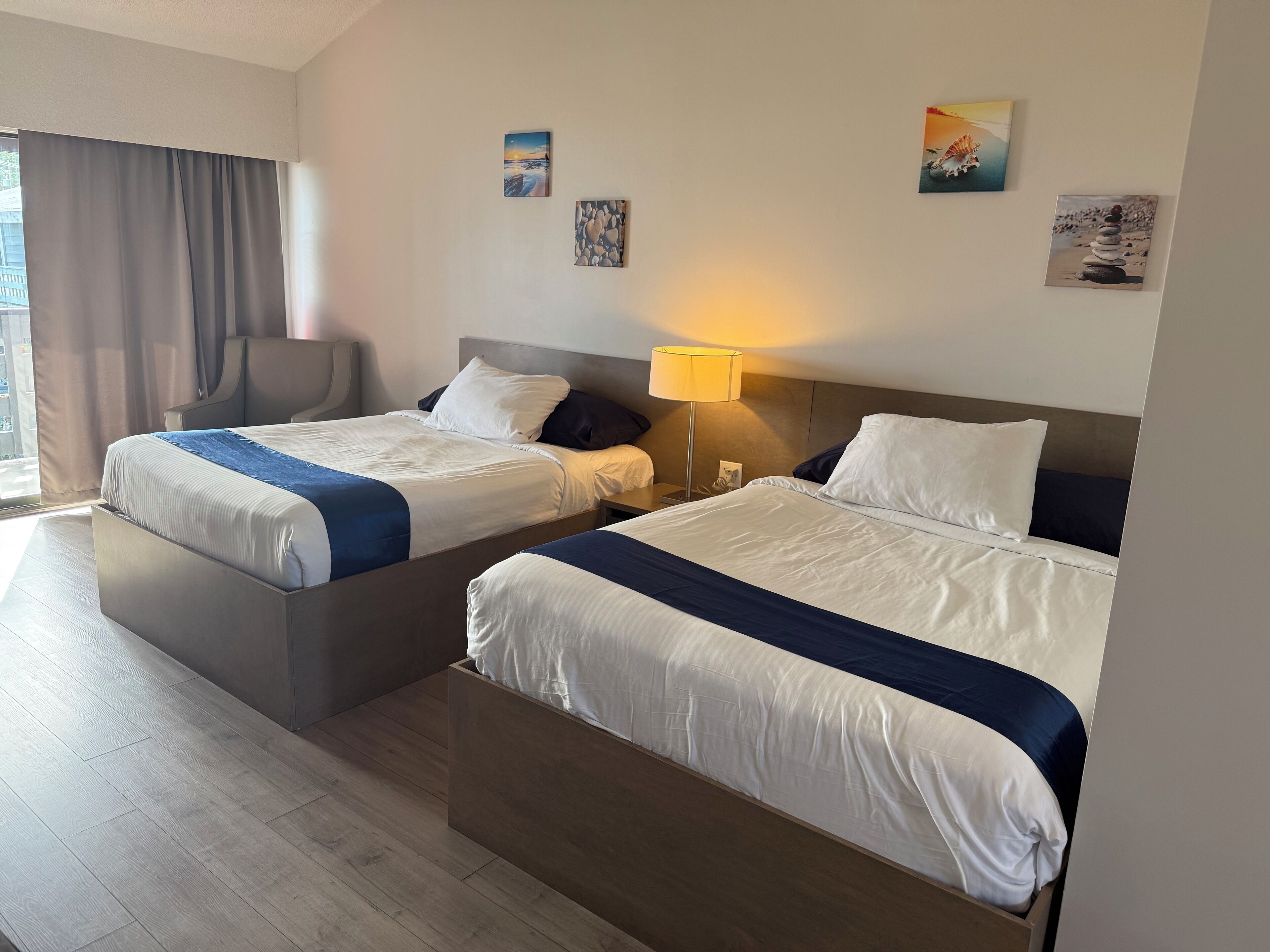 A hotel room with two beds, a desk, a chair, a lamp, and artwork on the walls.