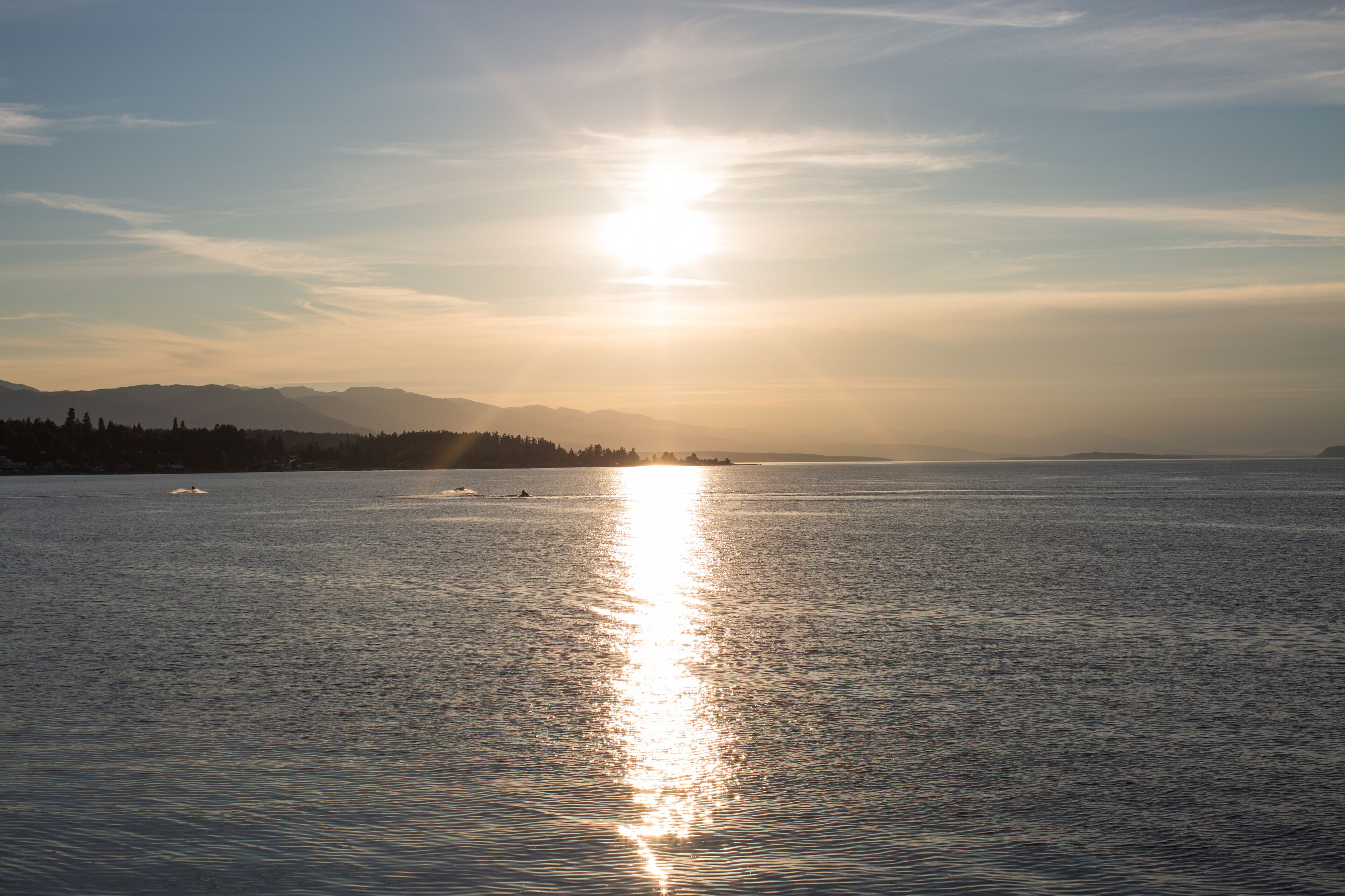 Een rustig waterlichaam met een felle zon die in het oppervlak spiegelt, omringd door een bebost landschap en verre bergen.